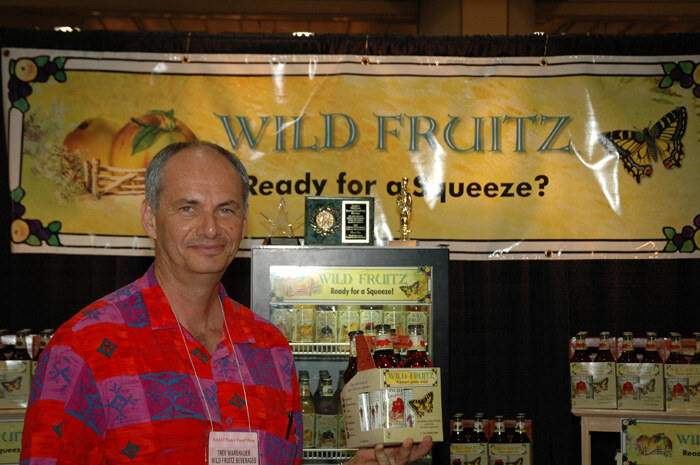 Trev Warshauer of Wild Fruitz Beverages, in a red patterned shirt, stands before a Wild Fruitz banner reading "Ready for a Squeeze?" and holds a carton of bottles near a display cooler.