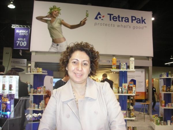 A smiling woman with curly hair stands in front of the Tetra Pak display, with shelves of packaged products and a Zola carton at left.