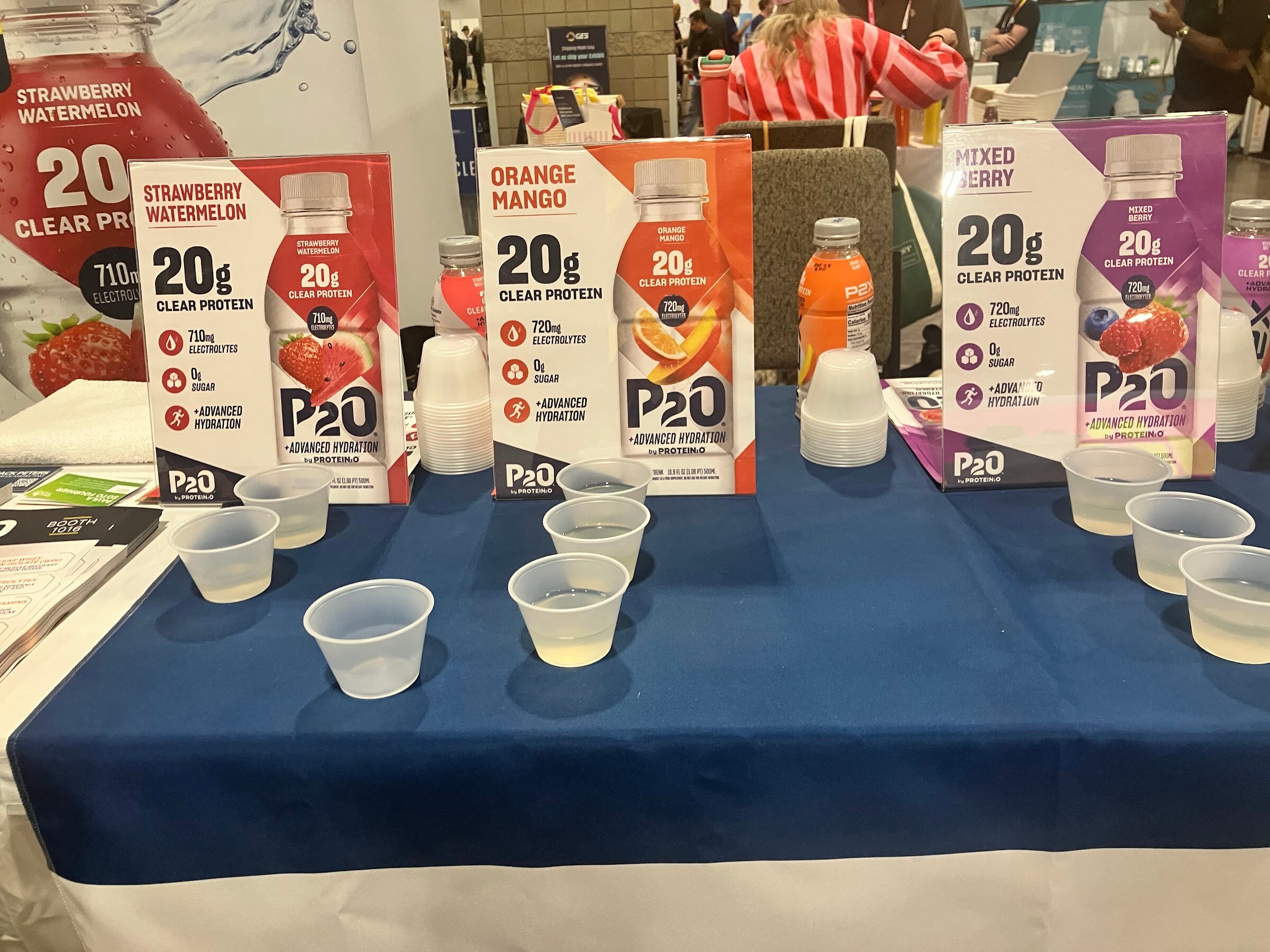 Sample cups on a blue tablecloth in front of P2O signs for strawberry watermelon, orange mango and mixed berry.