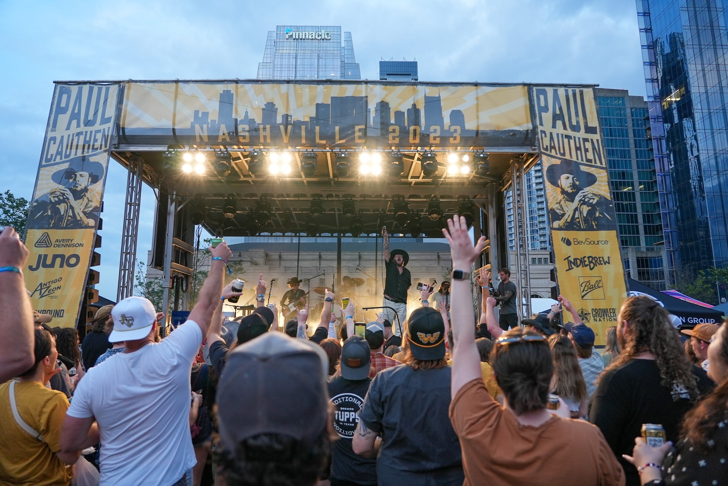 Fans with raised arms face a stage under a Paul Cauthen Nashville 2023 banner with downtown skyscrapers behind at dusk