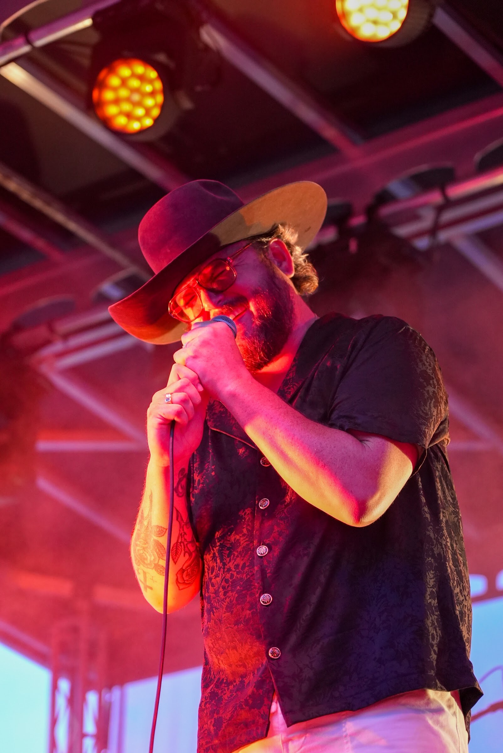 A singer in a wide-brim hat and dark patterned shirt holds a microphone to his mouth under red stage light