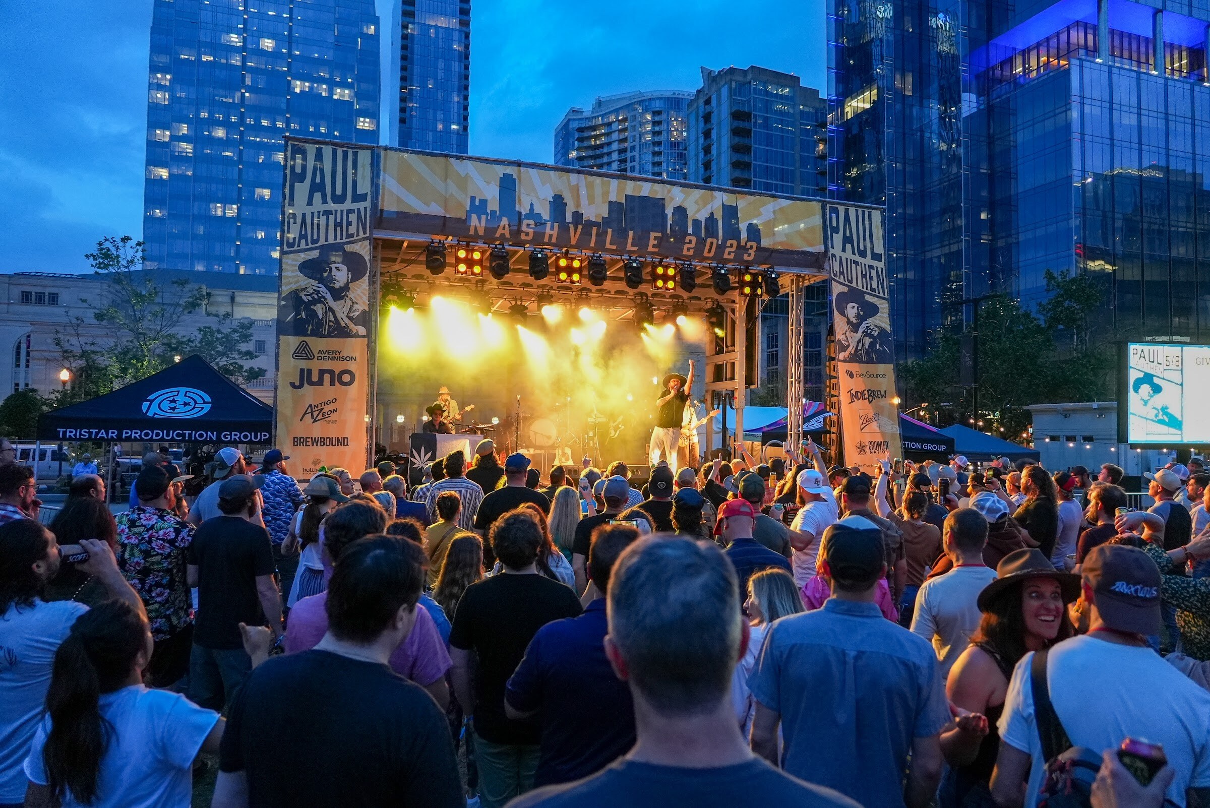 A crowd stands facing a lit stage framed by tall Paul Cauthen banners with sponsor logos, including Brewbound, and city towers in the background