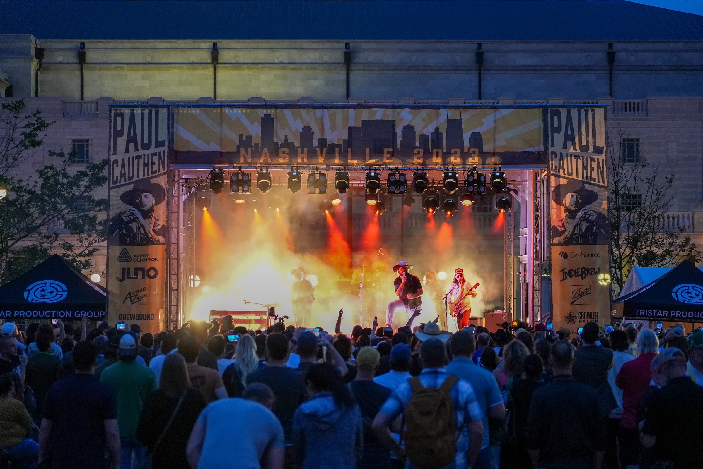 A band plays under orange stage lights in front of a Paul Cauthen Nashville 2023 backdrop with a large crowd in the foreground