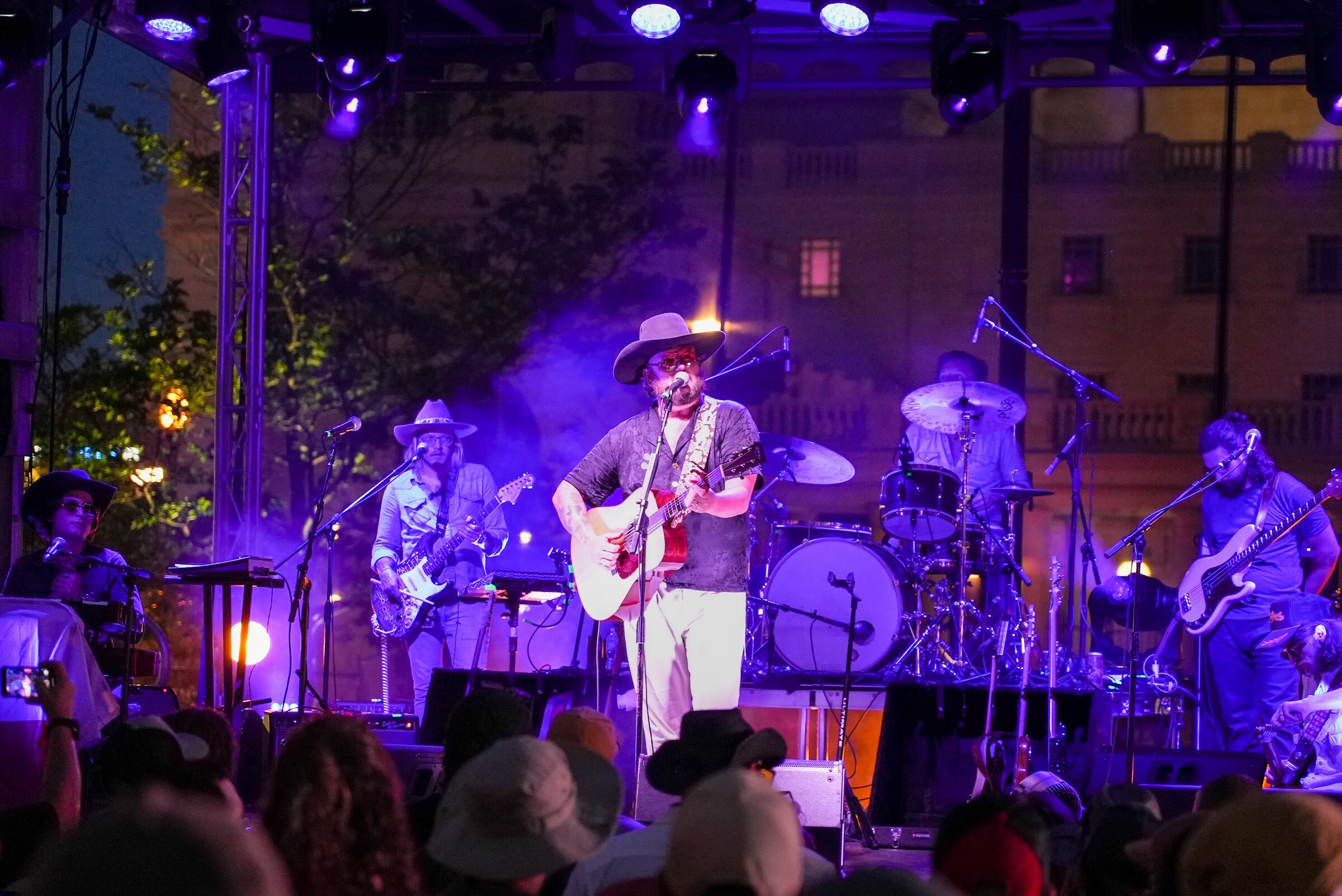 A guitarist in a cowboy hat sings at center stage with a drummer and band members behind him under purple lighting and haze