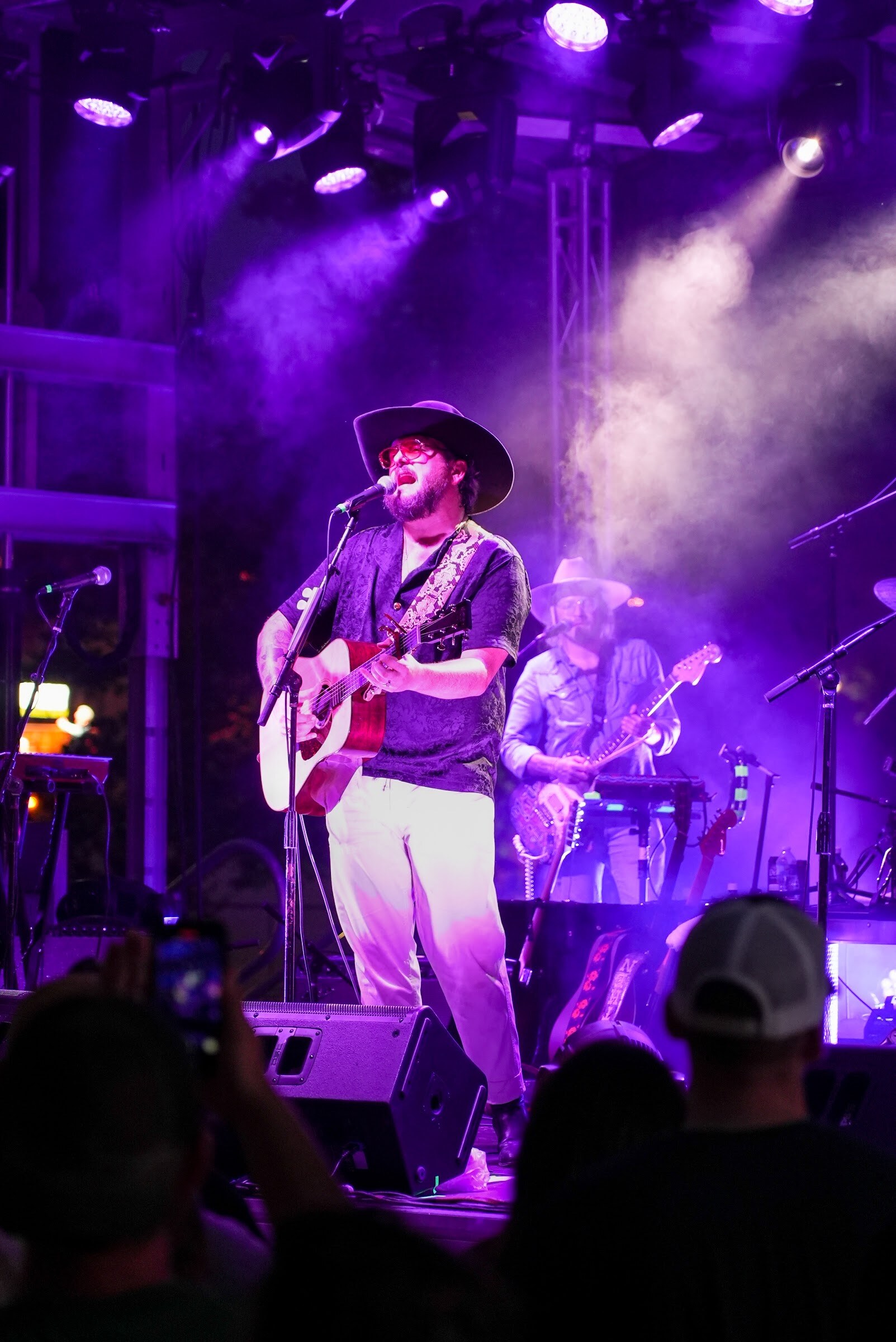 A singer in a cowboy hat plays an acoustic guitar on stage lit in purple with smoke behind him