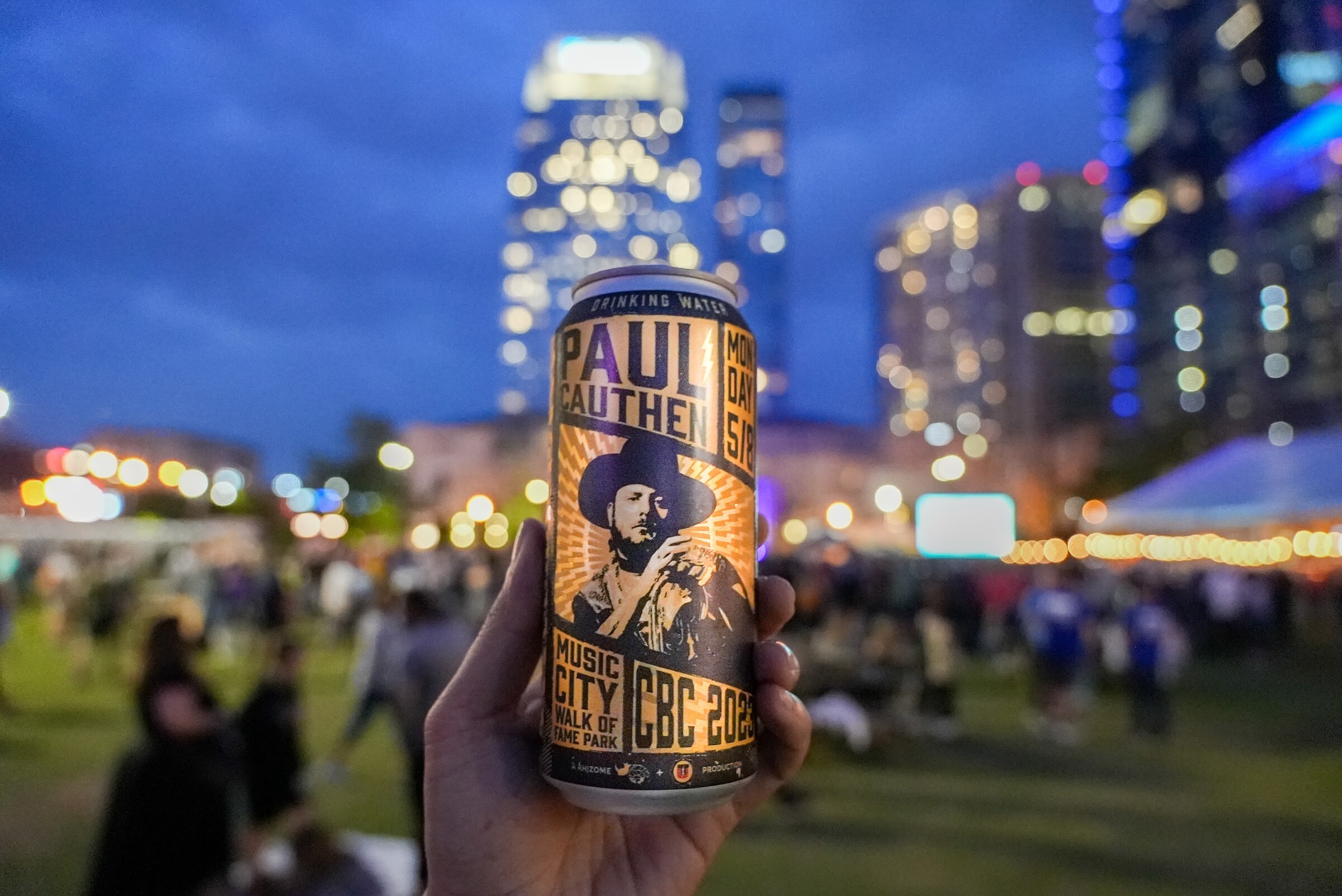 A hand holds a Paul Cauthen Music City Walk of Fame Park CBC 2023 commemorative can with blurred city lights behind it