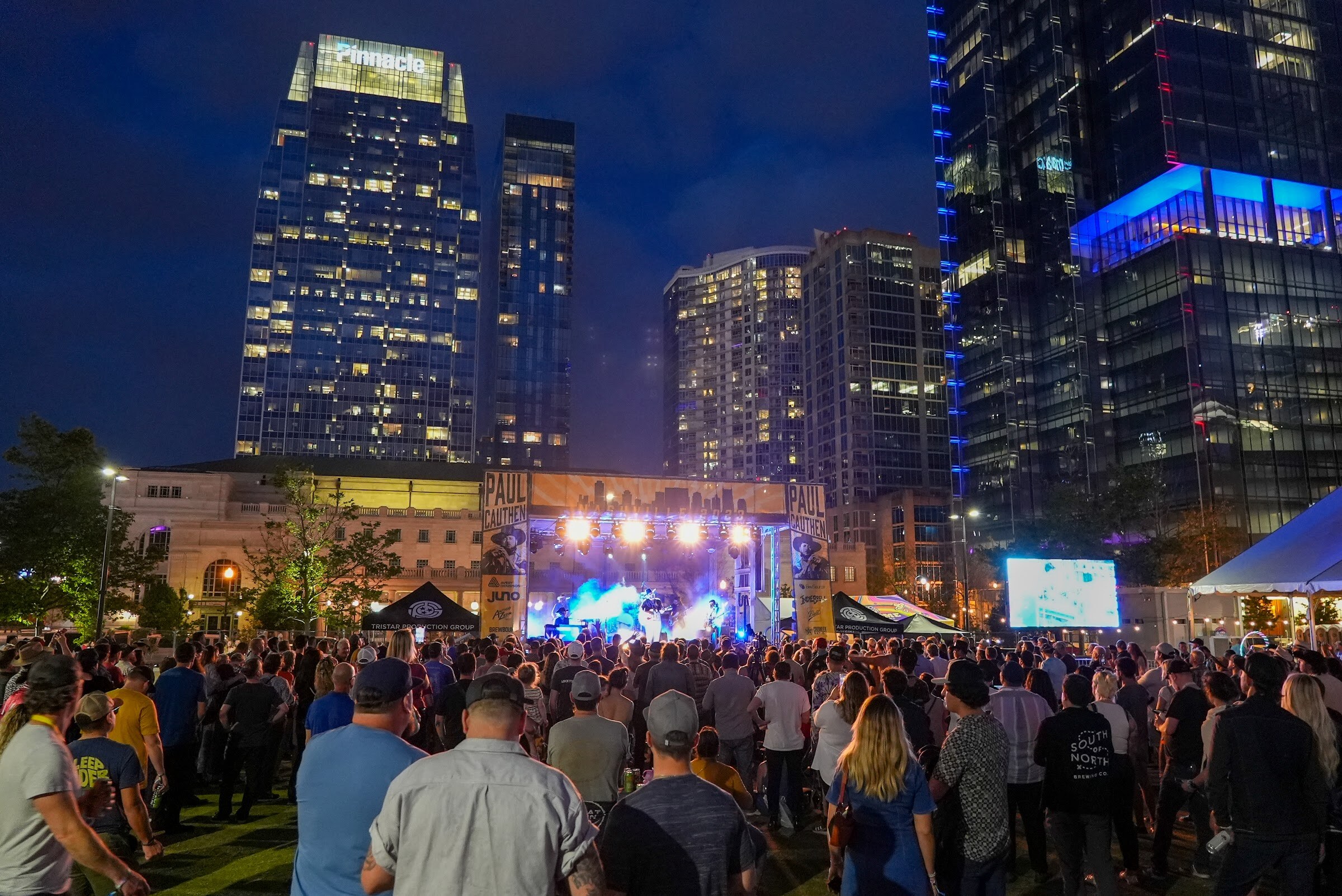 A packed lawn of attendees faces a lit stage with downtown towers glowing overhead