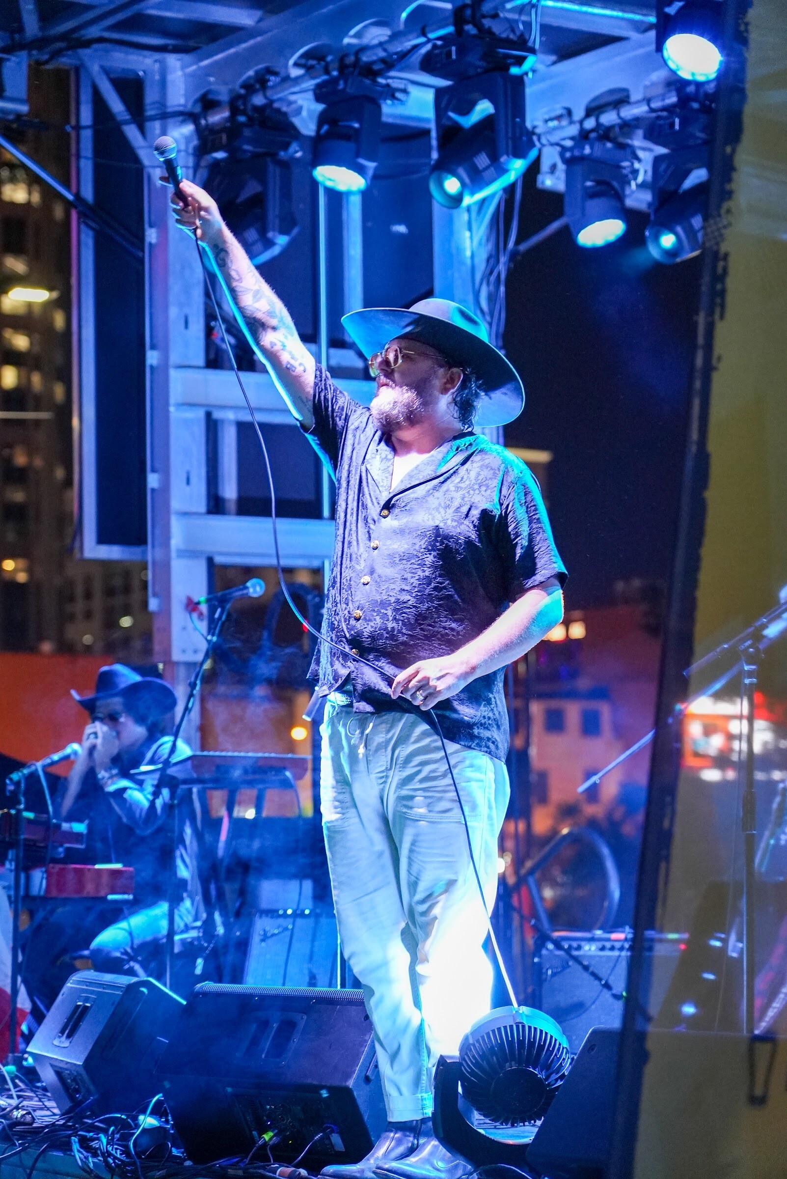 A singer in a cowboy hat points upward on stage lit in blue with city buildings in the background