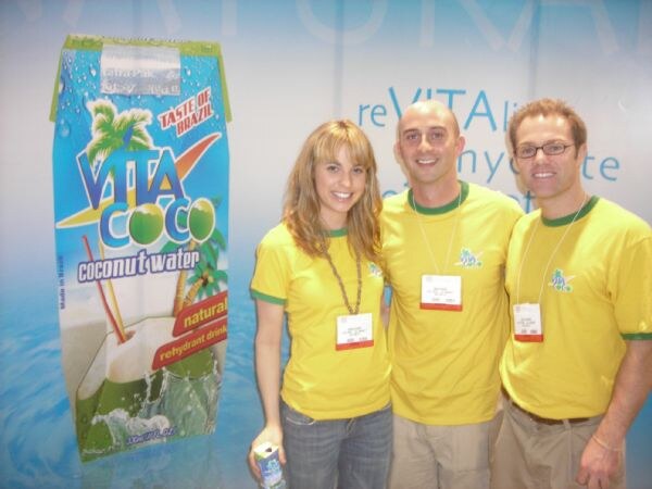 A woman and two men in yellow shirts with green trim stand beside a large Vita Coco coconut water carton graphic. The woman holds a small carton.