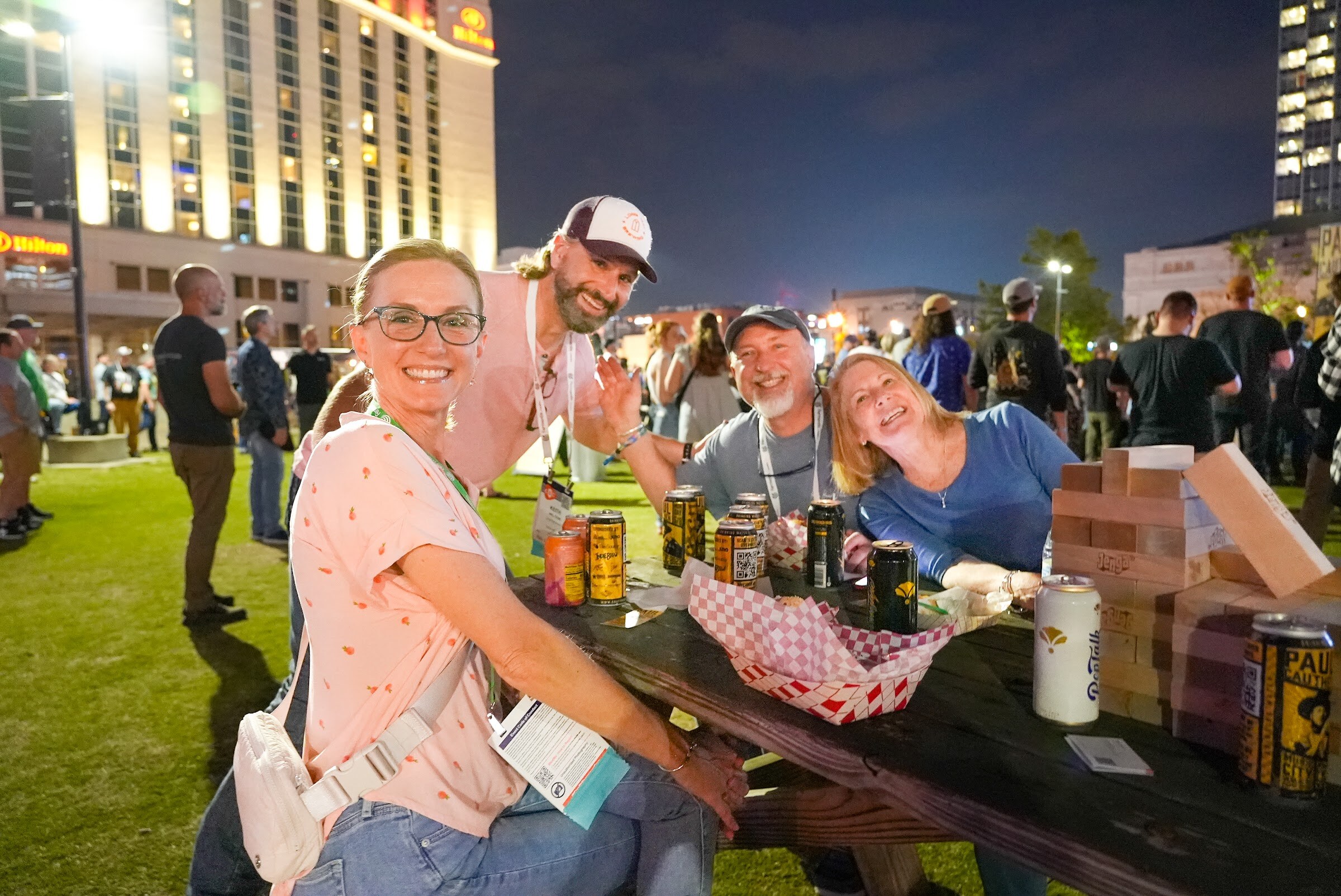 Four attendees with conference badges pose at a picnic table with cans, a basket and wooden blocks on the lawn at night