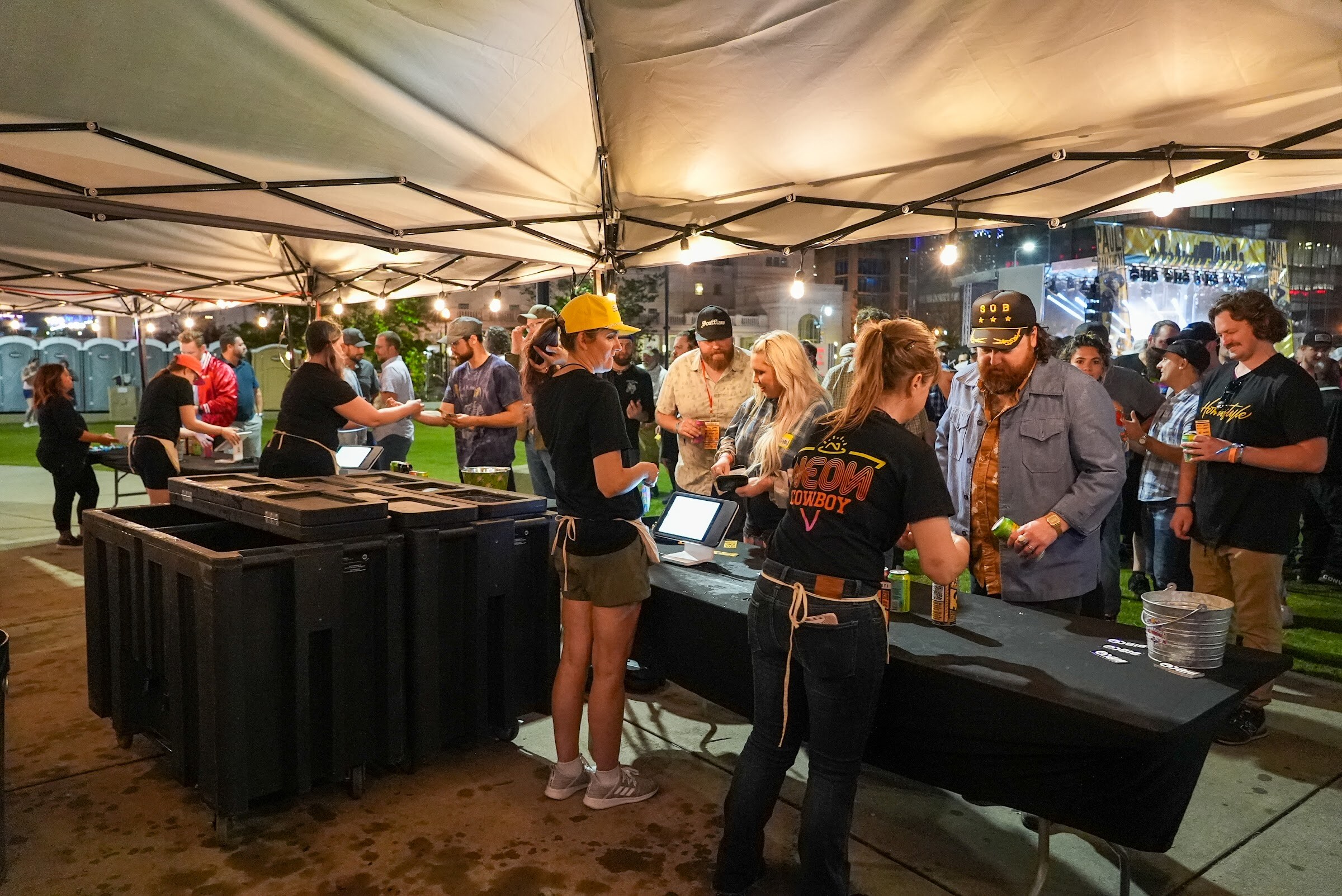 Staff in black shirts serve beer from a long table under a white tent while attendees wait and string lights glow overhead