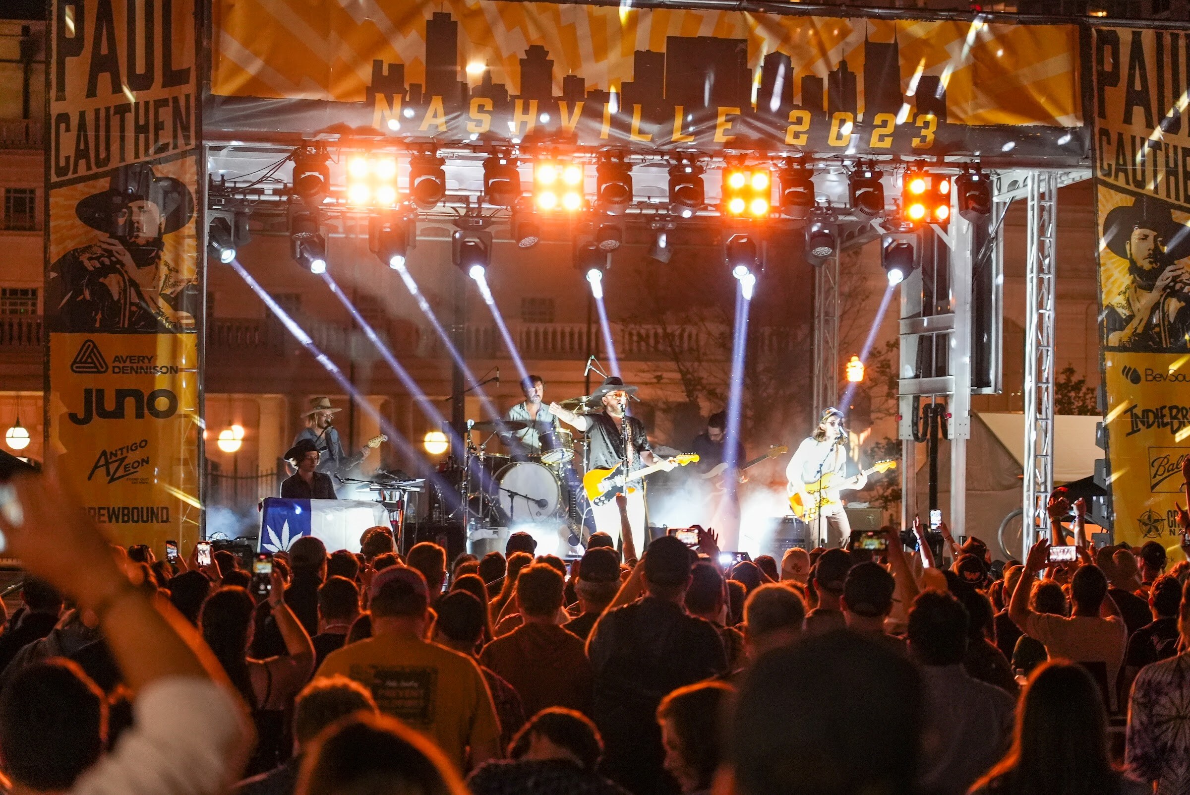 A band plays on a stage with Paul Cauthen banners and sponsor logos while spotlight beams cut through haze above the crowd