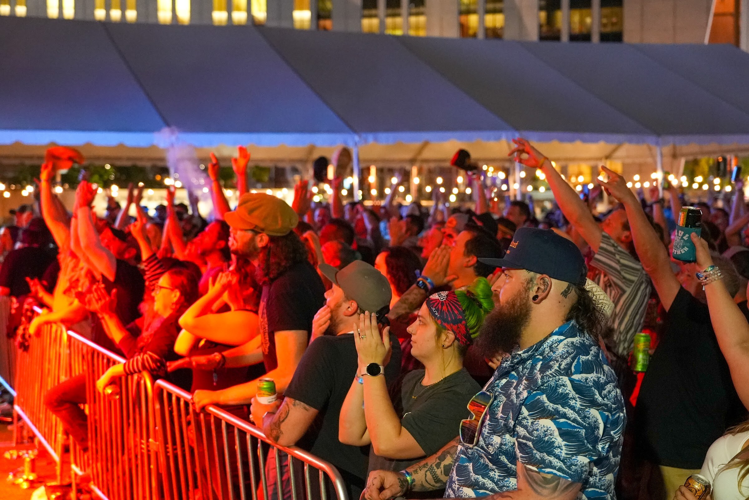 Fans in hats and Hawaiian shirts crowd a metal barrier with raised arms under red light and a tent canopy