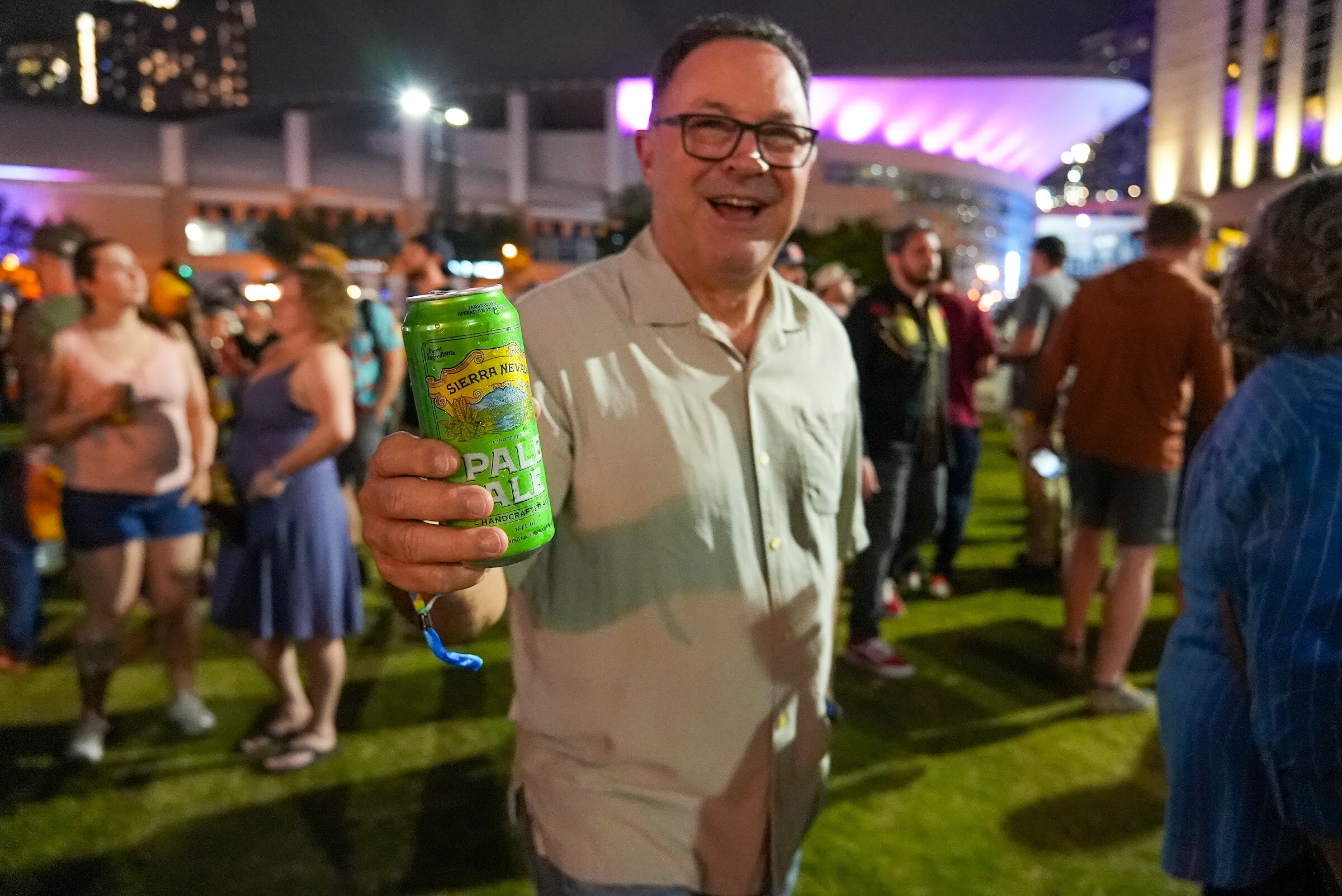 A smiling man in glasses holds up a green Sierra Nevada Pale Ale can on the lawn with other attendees behind him