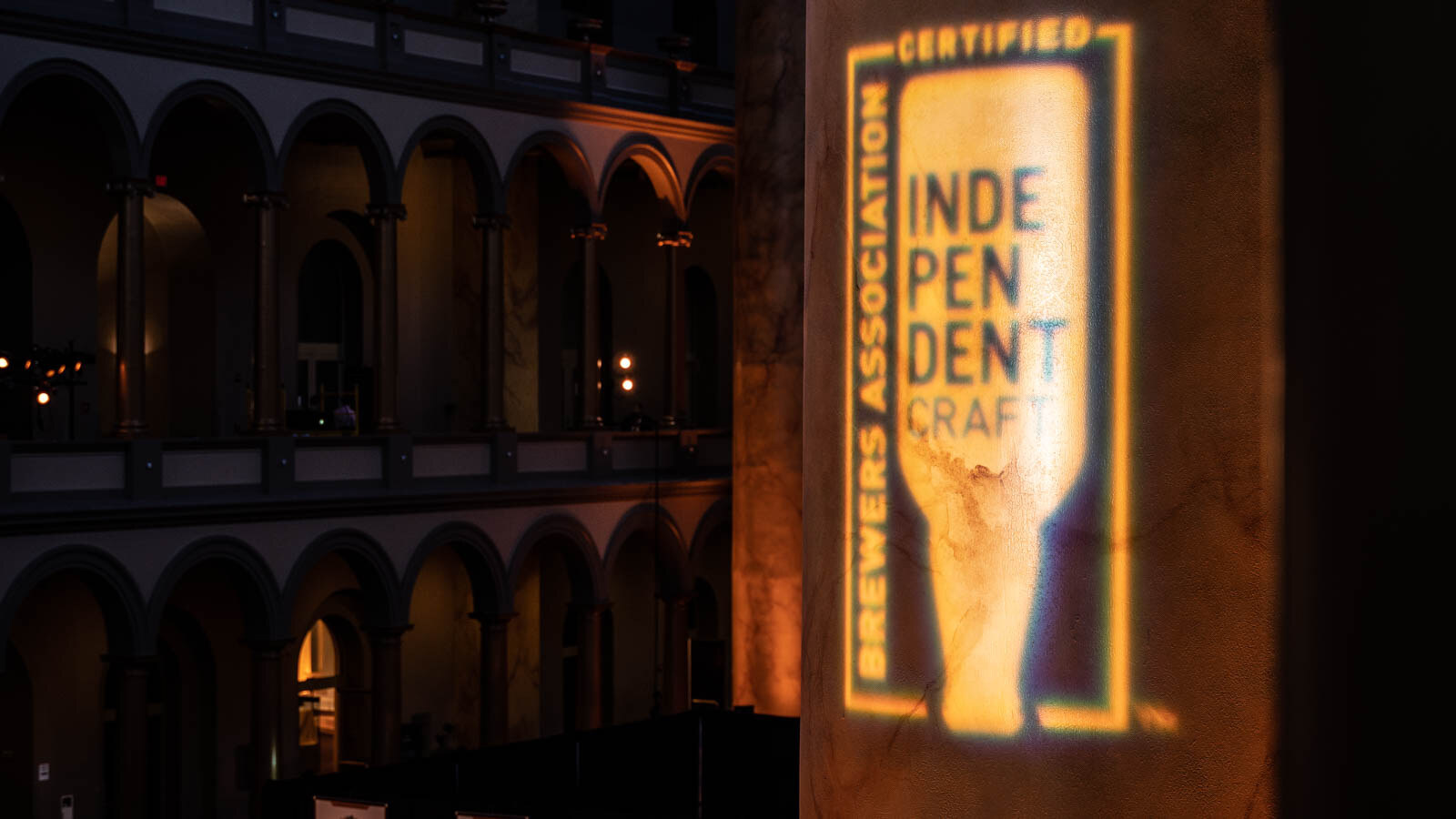 A projected Brewers Association Certified Independent Craft logo on a stone column, with arched balconies in the background.