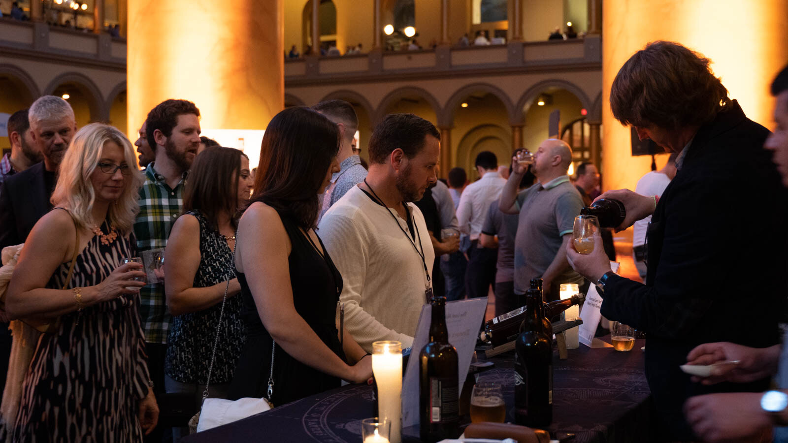 Attendees standing in line at a draped table with bottles and a candle, a man at the right pouring a glass.