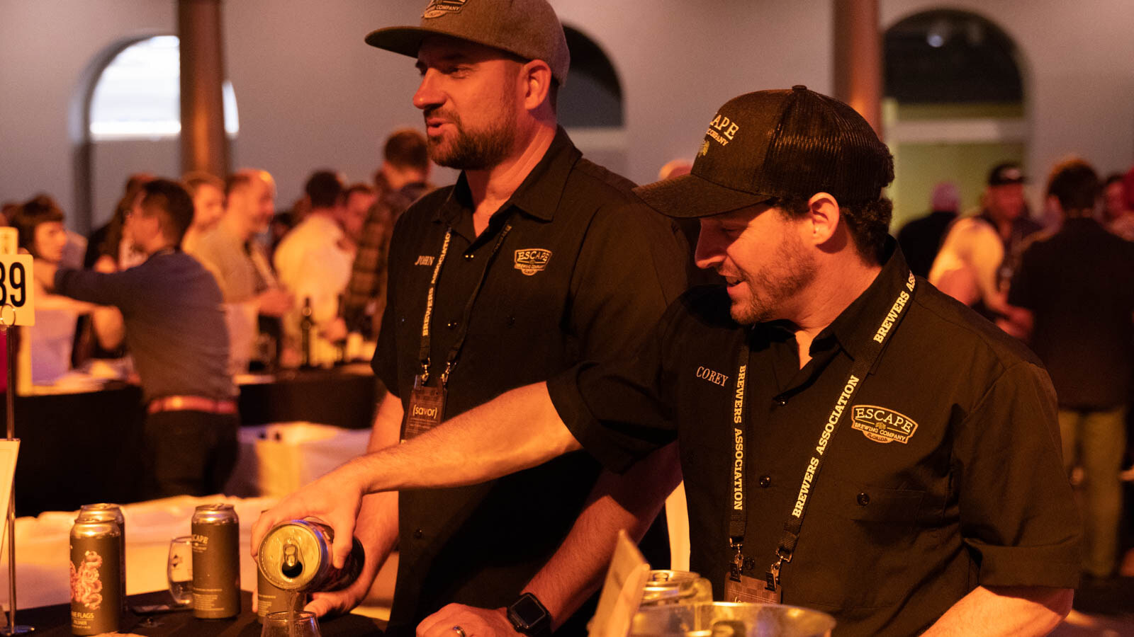 Two men in black work shirts and caps with lanyards pouring beer at a table with cans.
