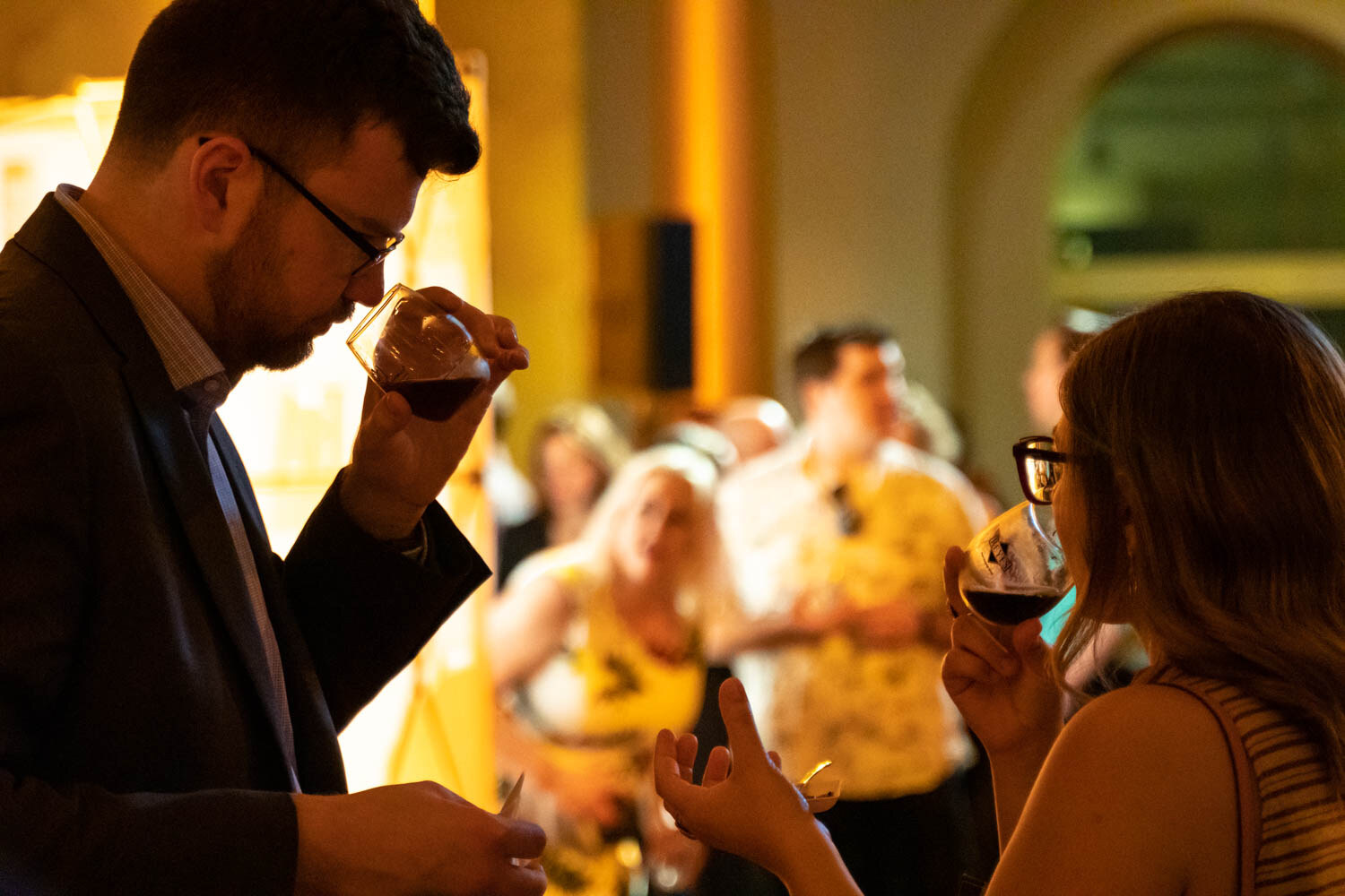 A bearded man in glasses and a blazer holding a glass of dark beer to his nose next to a woman drinking from her glass.