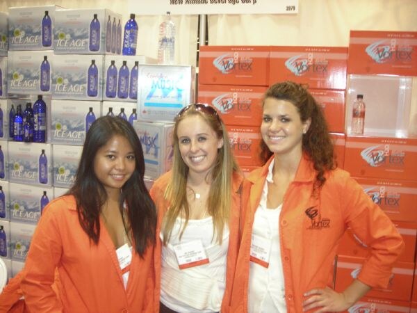 Three smiling women in orange Vortex jackets stand in front of stacked cases of Ice Age and Vortex bottled water.