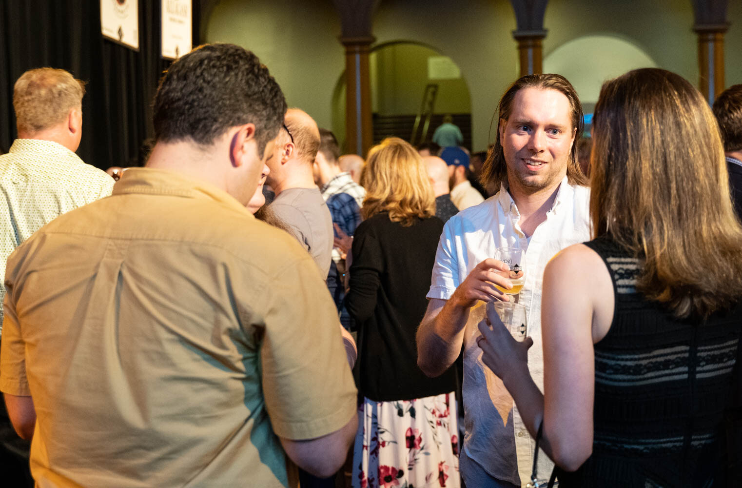 A small group of attendees talking and holding glasses of beer, with draped tables and arches behind them.