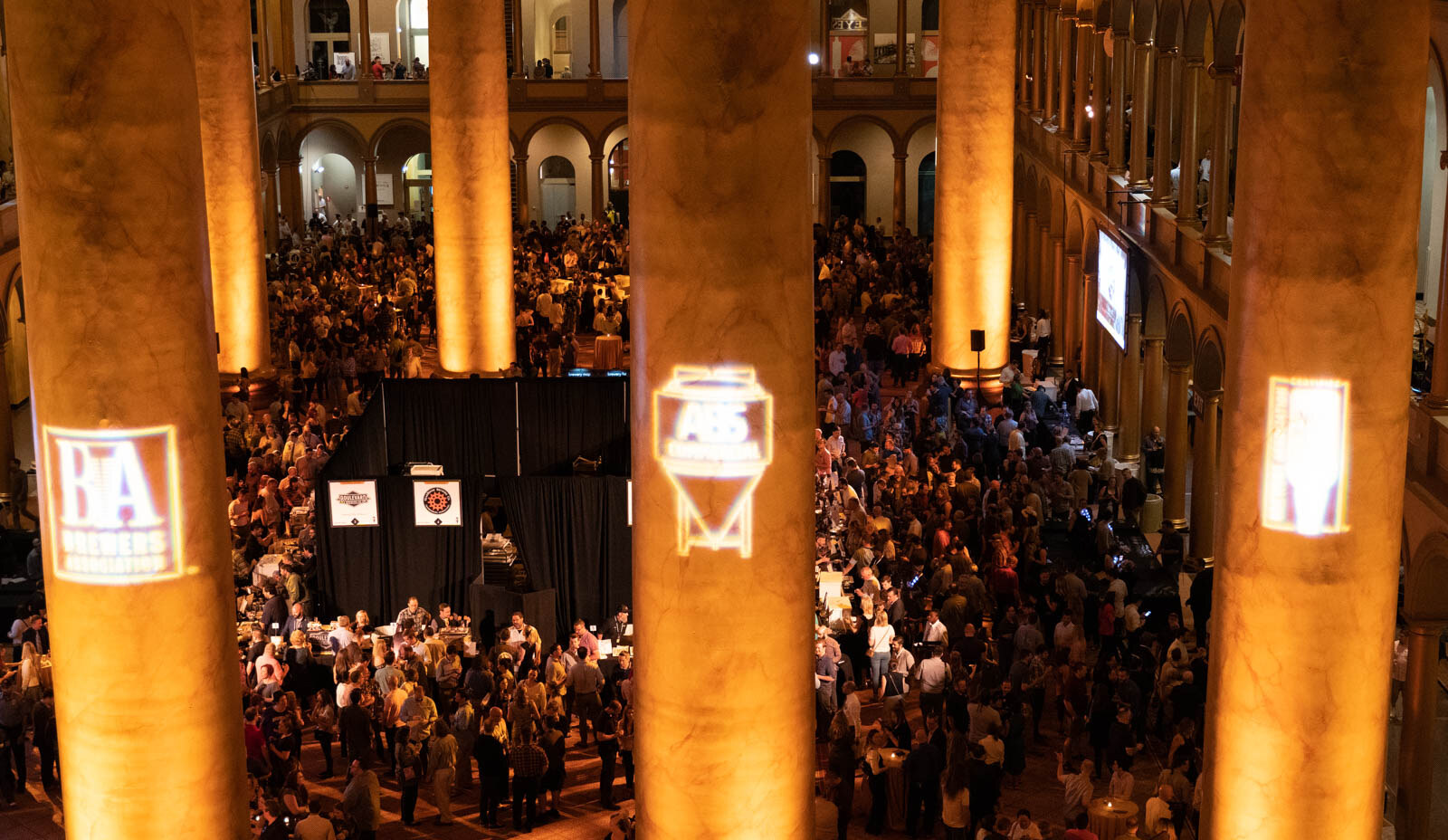 View between stone columns carrying glowing projected logos, down to a crowd of attendees below.