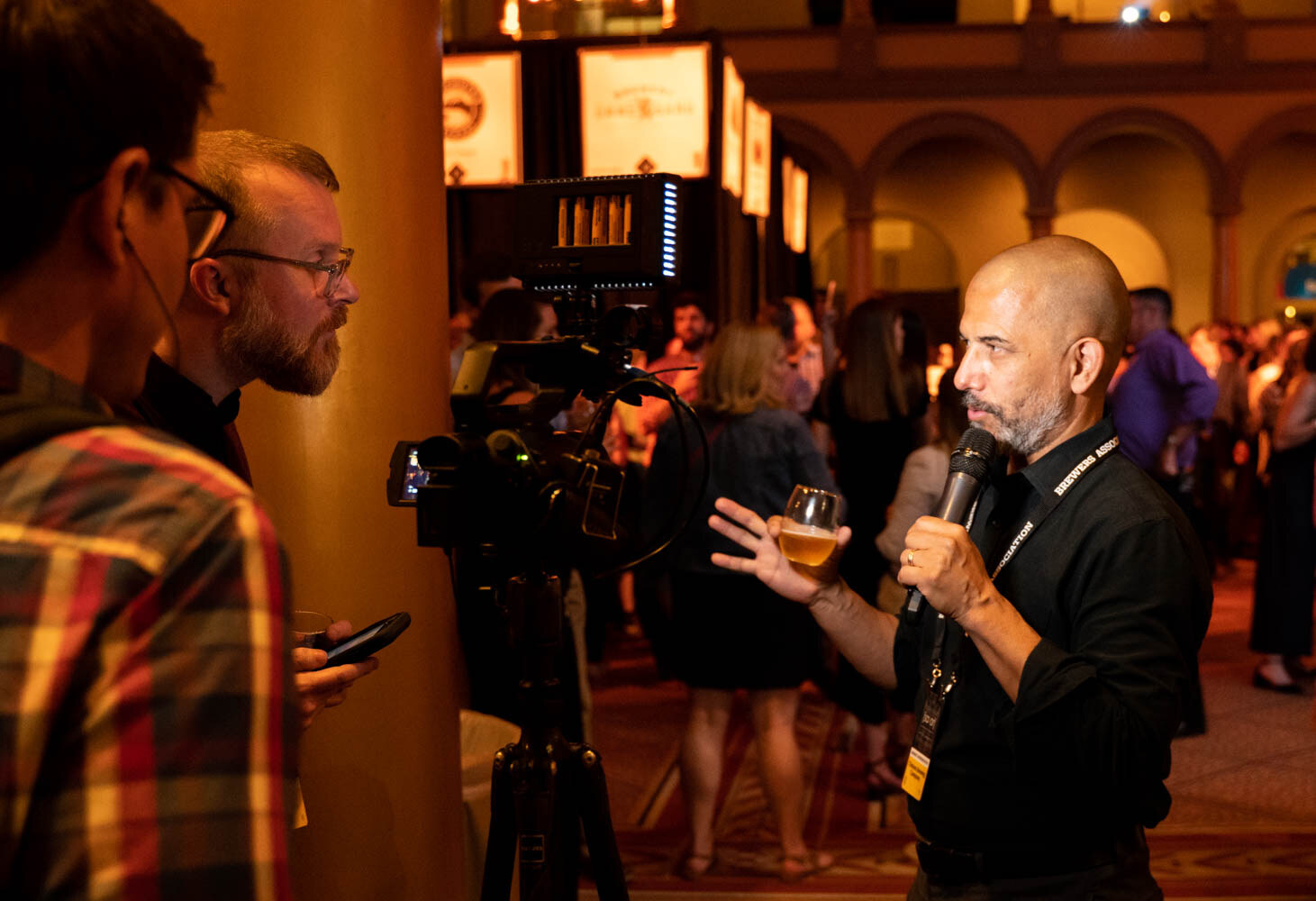 A man with a lanyard, a glass of beer and a microphone talking to two people and a video camera on a tripod.