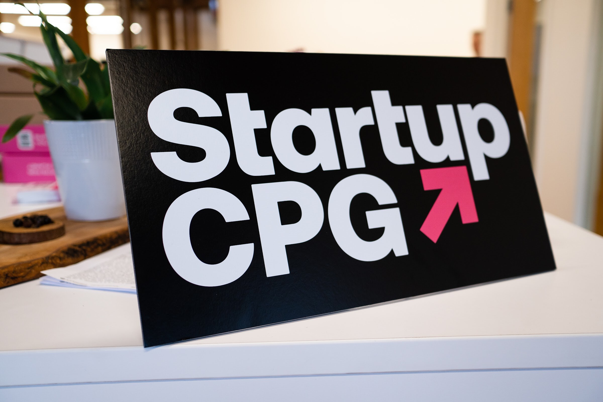A black and white Startup CPG sign with a pink arrow sits on a white table beside a plant
