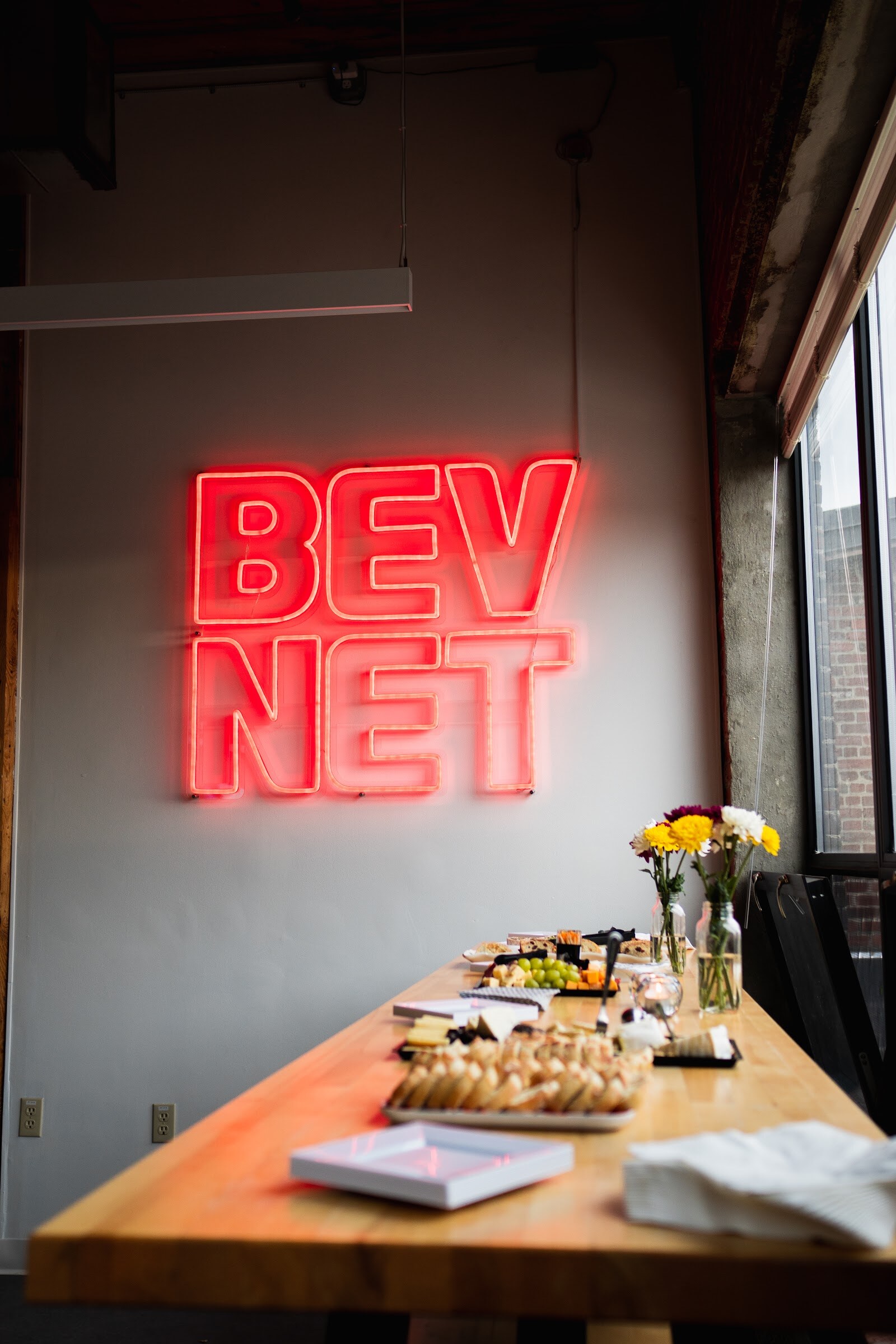 A red BEVNET neon sign glows on a wall above a wooden table set with flowers and snacks
