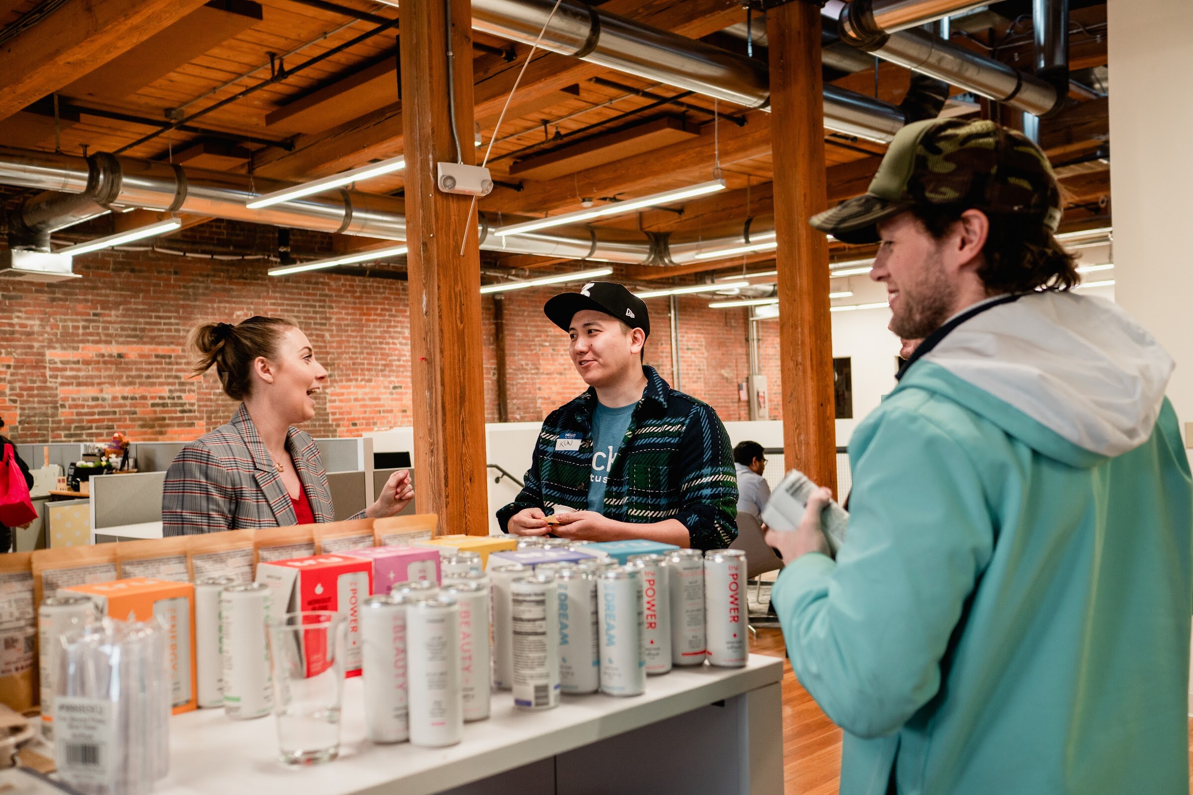 A woman in a plaid blazer and a man in a plaid shirt chat with a guest in a teal jacket across a table lined with tall slim cans and boxed products