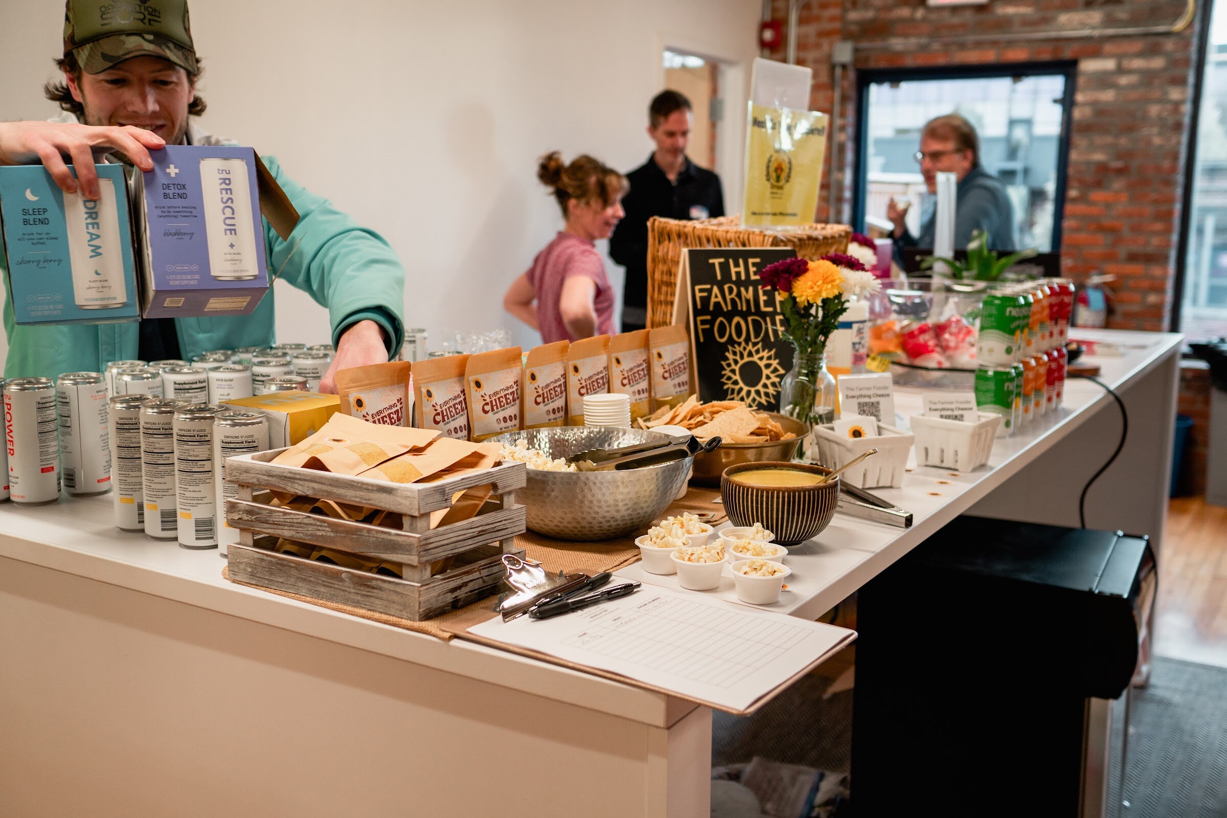 A host reaches for boxed drinks on a long table with chips, cheese snacks, a Farmer Foodie chalkboard sign and cans