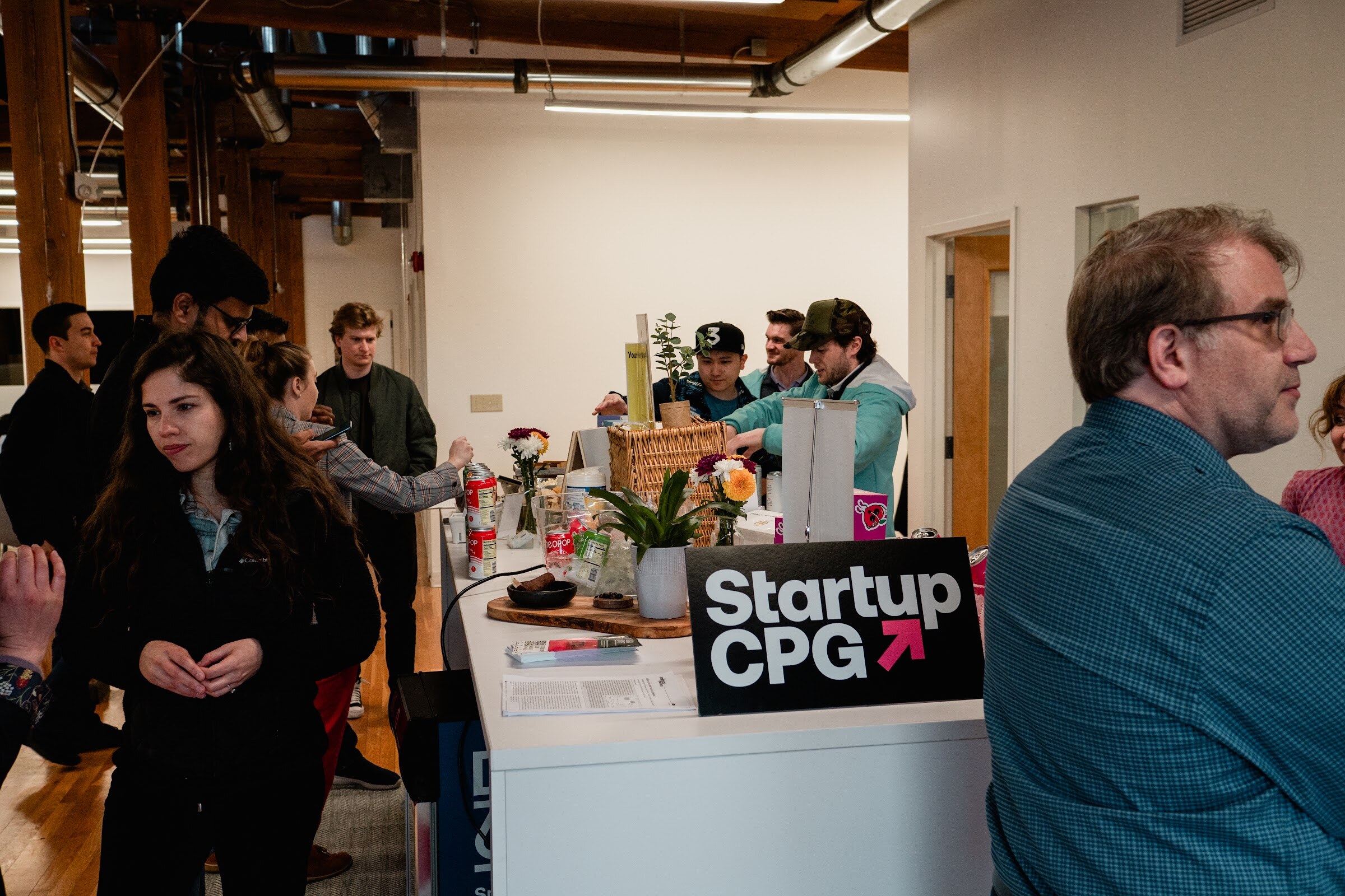 Guests stand around a white table with a Startup CPG sign and products in a brick and timber loft office