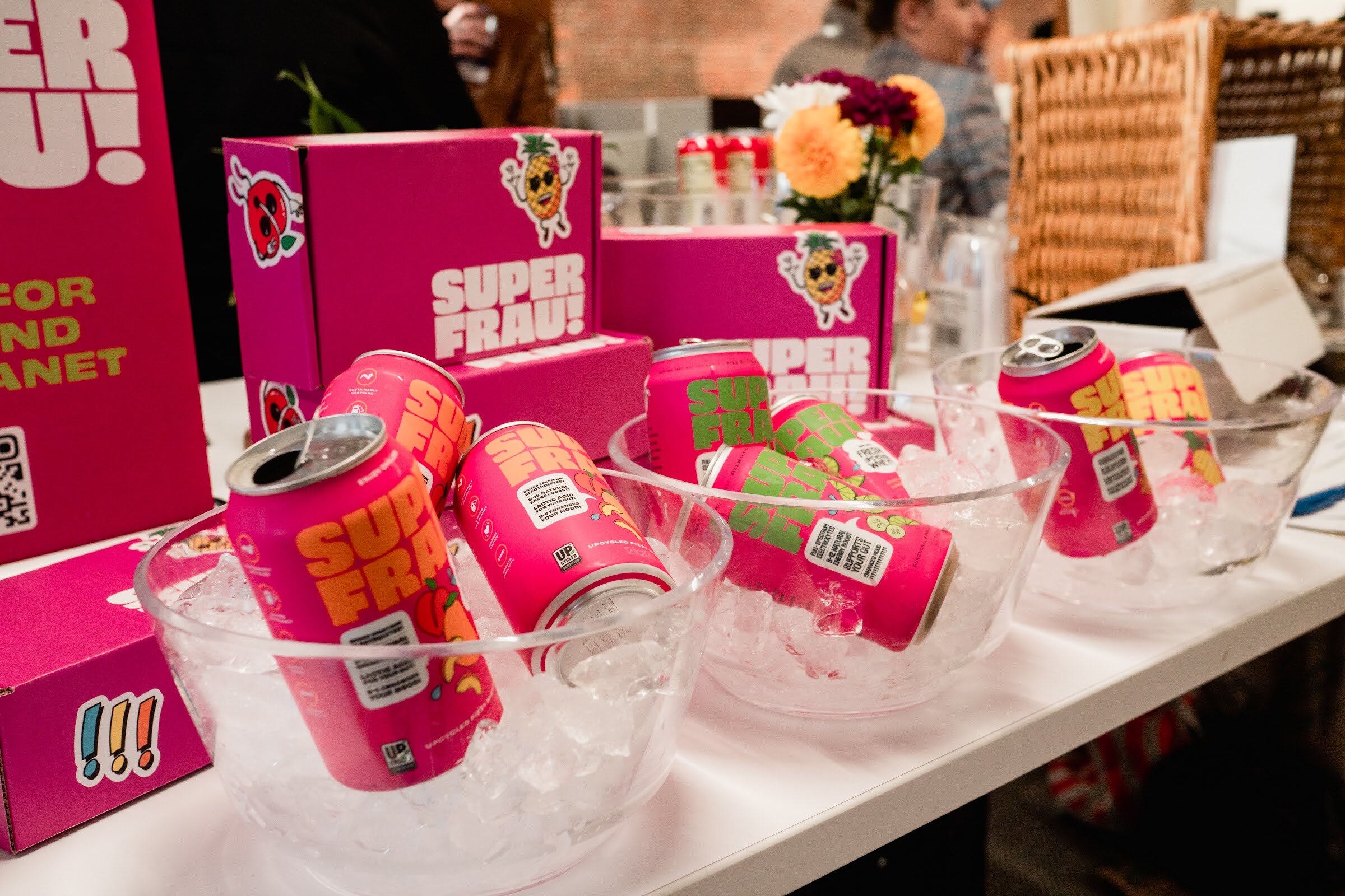 Pink Super Frau cans sit in clear bowls of ice beside pink Super Frau boxes and a bouquet of flowers