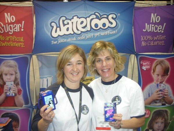 Two smiling women in white Wateroos shirts hold small Wateroos drink packages in front of a blue Wateroos banner and panels reading No Sugar and No Juice.