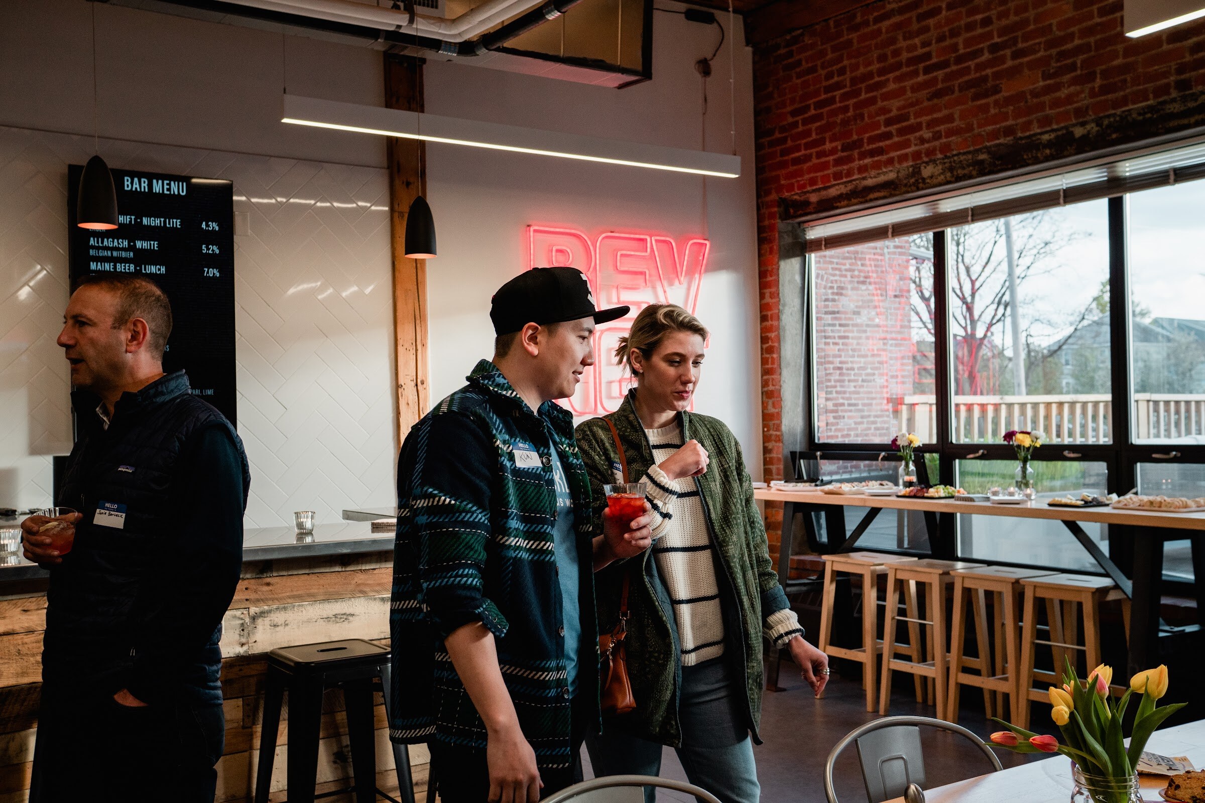 Attendees with name tags talk near a bar menu board, a red neon BEVNET sign and a window overlooking brick buildings