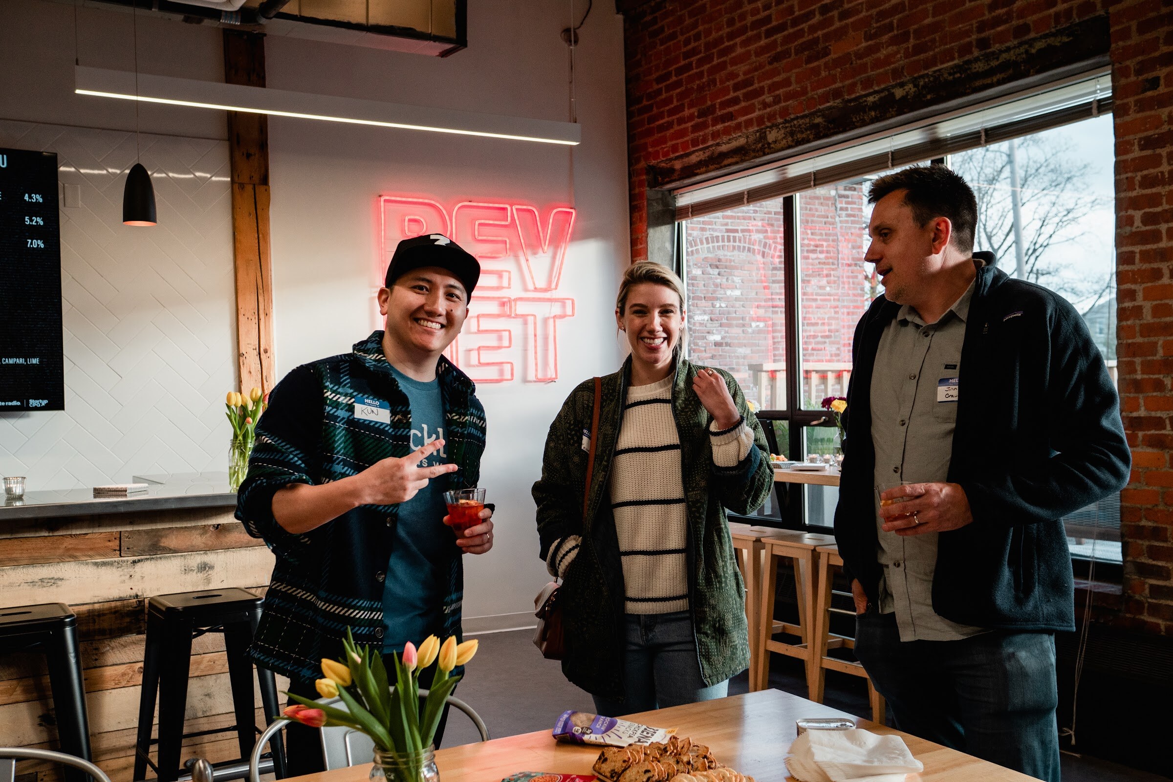 A man throws a peace sign next to a smiling woman and a man holding a drink in front of a window and red neon sign
