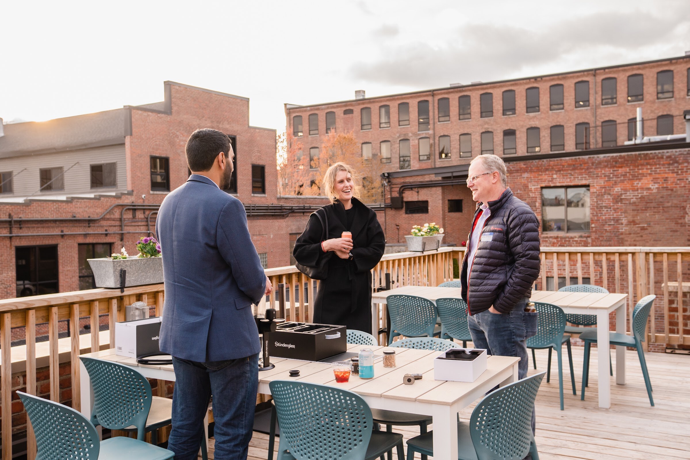 Three guests talk around a patio table on a wooden rooftop deck with brick mill buildings behind them