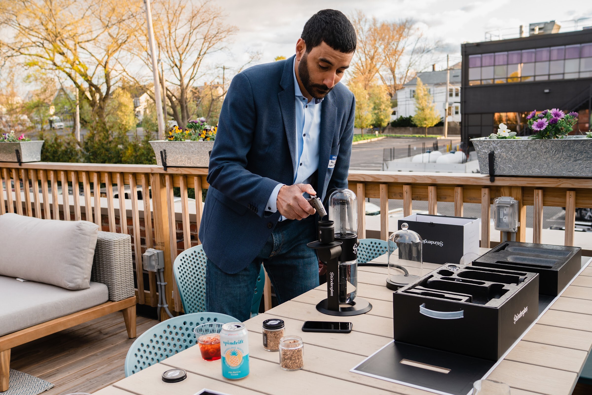 A man in a blue blazer works a coffee grinder on a deck table beside black cases, a canned drink and small jars
