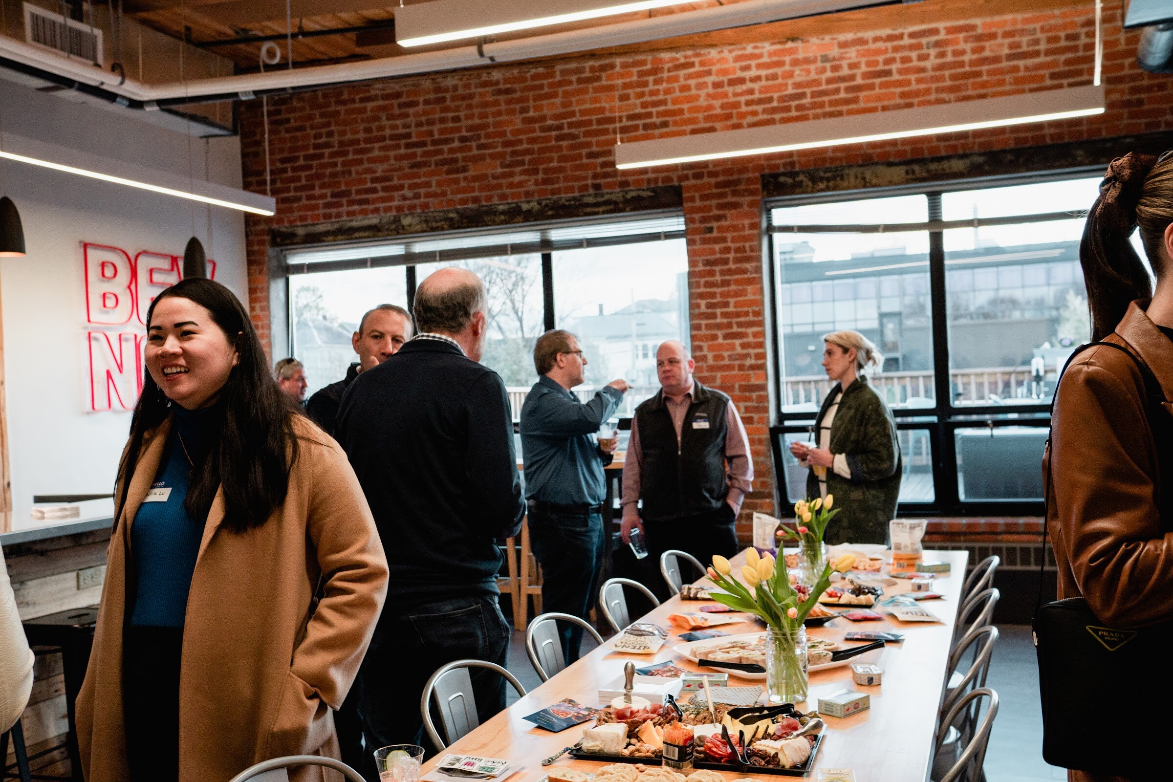 A smiling woman in a camel coat stands beside a long wooden table of snacks and tulips while other guests talk by the windows