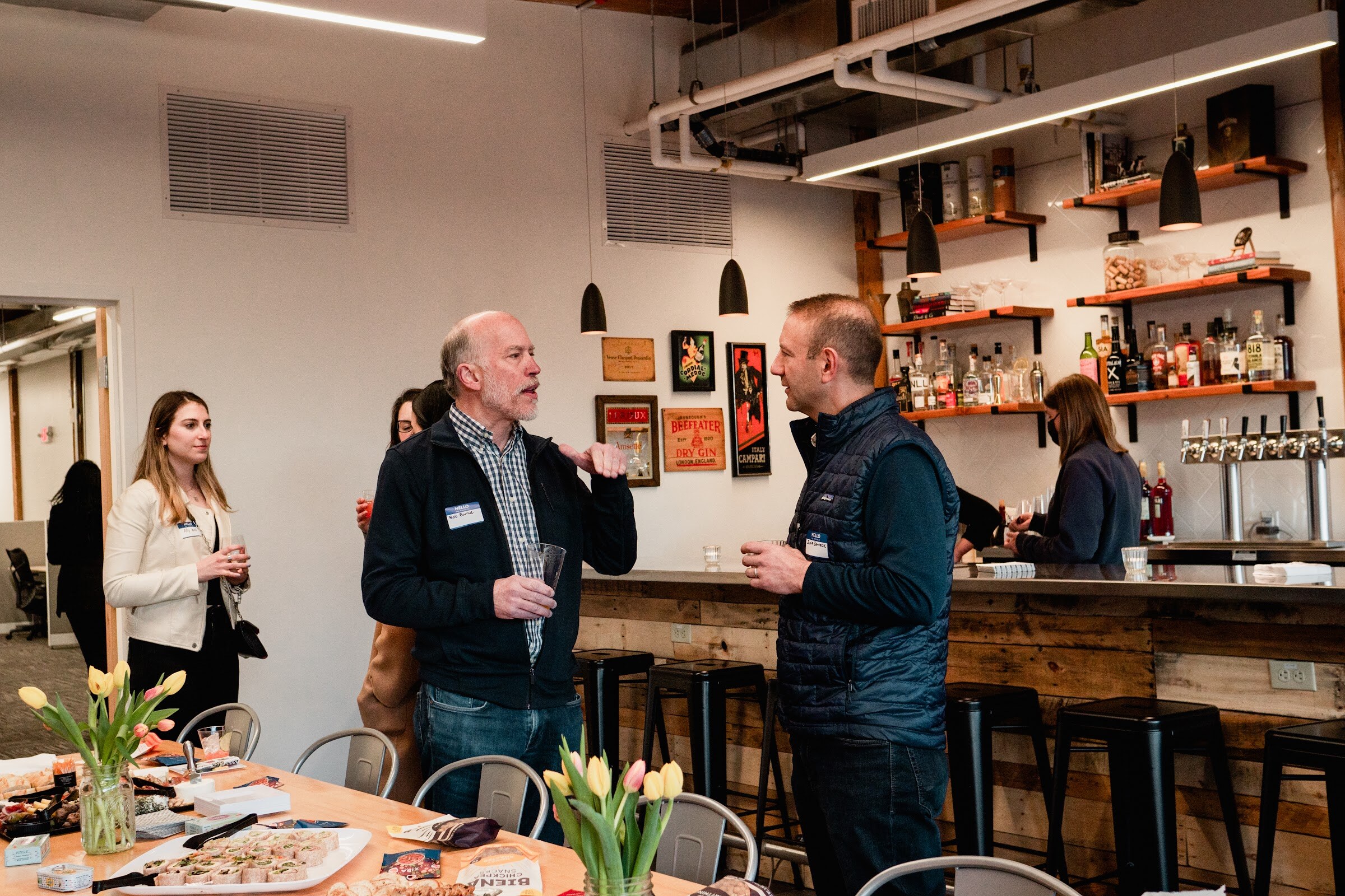 Two men with name tags talk near a long snack table and a bar with shelves of bottles and taps