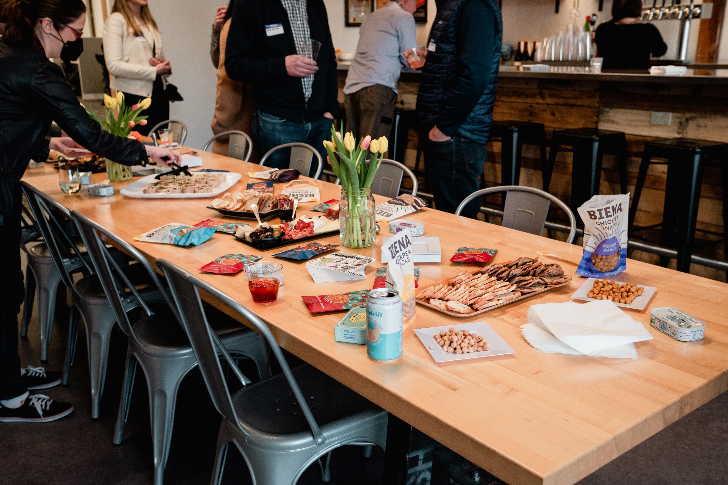 A long wooden table holds platters of charcuterie, bags of chickpea snacks, a tin of fish, a canned drink and a vase of tulips