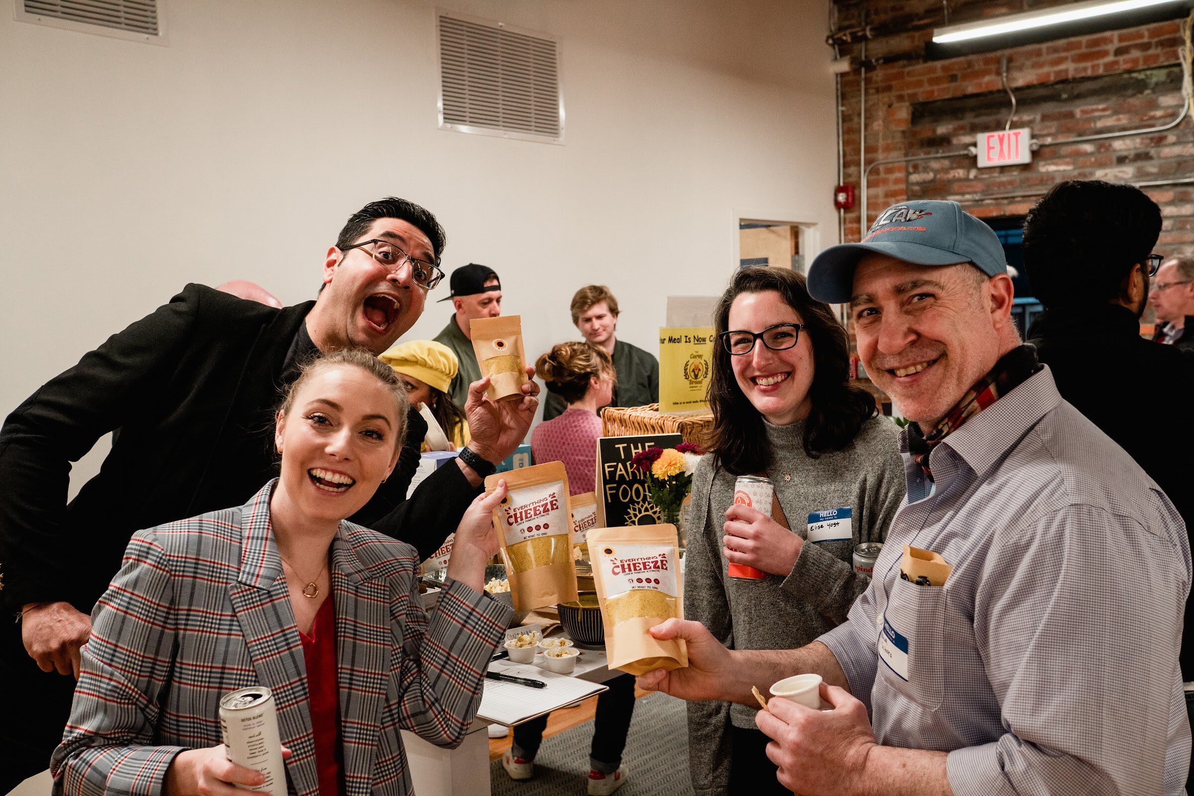 Four smiling attendees pose for the camera holding snack bags and cans at a sample table
