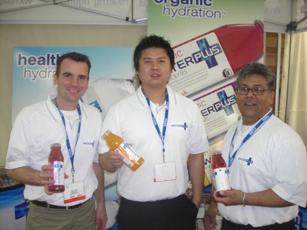 Three men in white Water Plus shirts and lanyards stand together, each holding a bottle of Water Plus, in front of organic hydration signs.