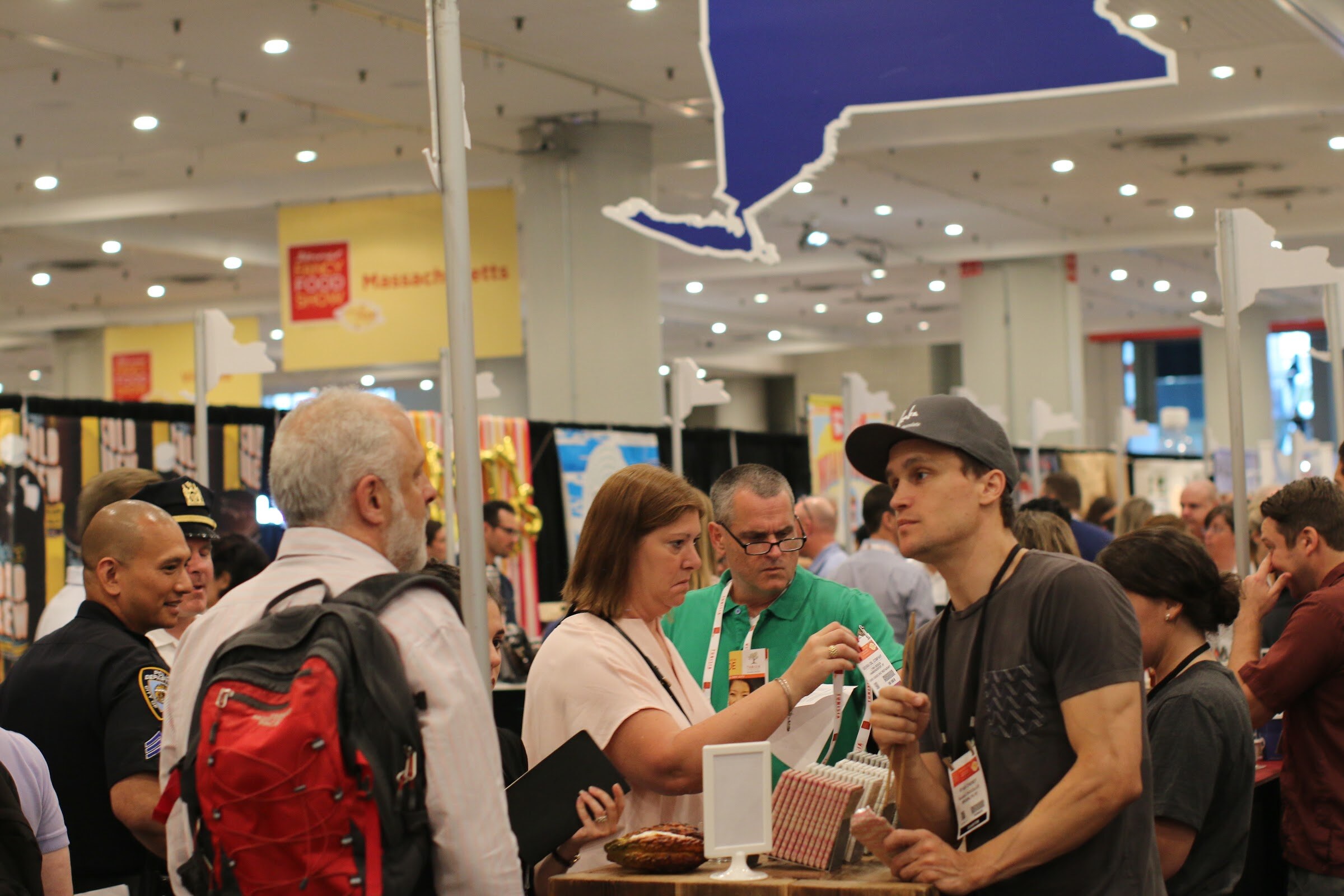 Group of badge-wearing attendees around a wooden counter with cacao pods, under a large blue New York-shaped sign.