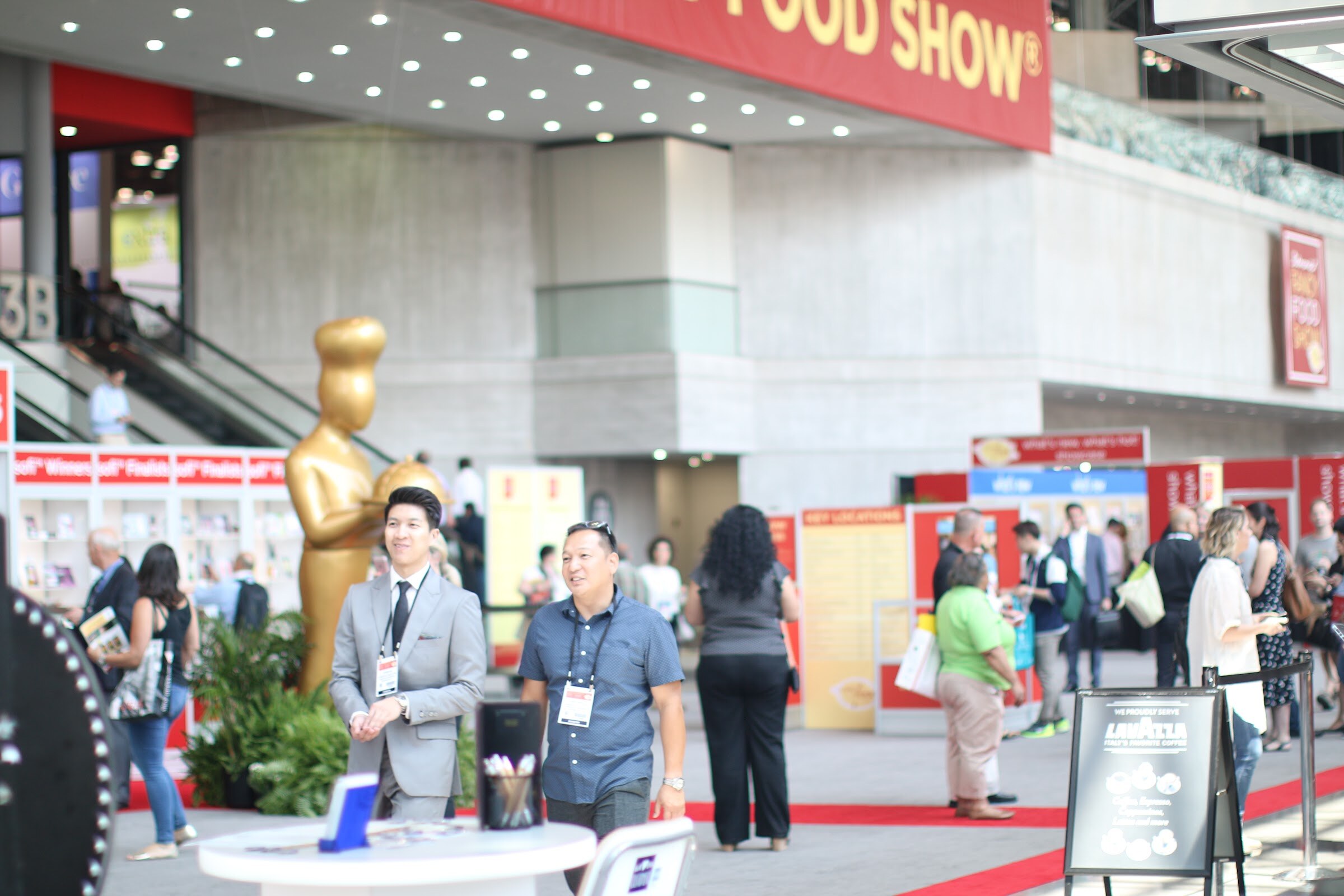 Two smiling men with badges at a round table in front of a tall gold statue, with attendees and red carpet behind them.