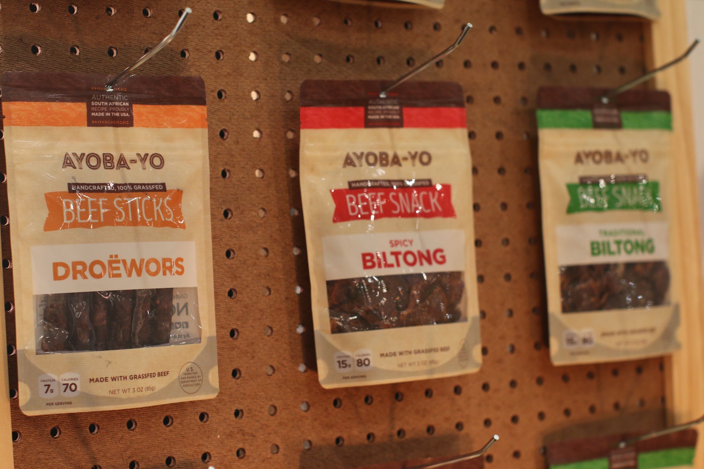 Three Ayoba-Yo pouches, droewors, spicy biltong and traditional biltong, hanging from hooks on a pegboard.
