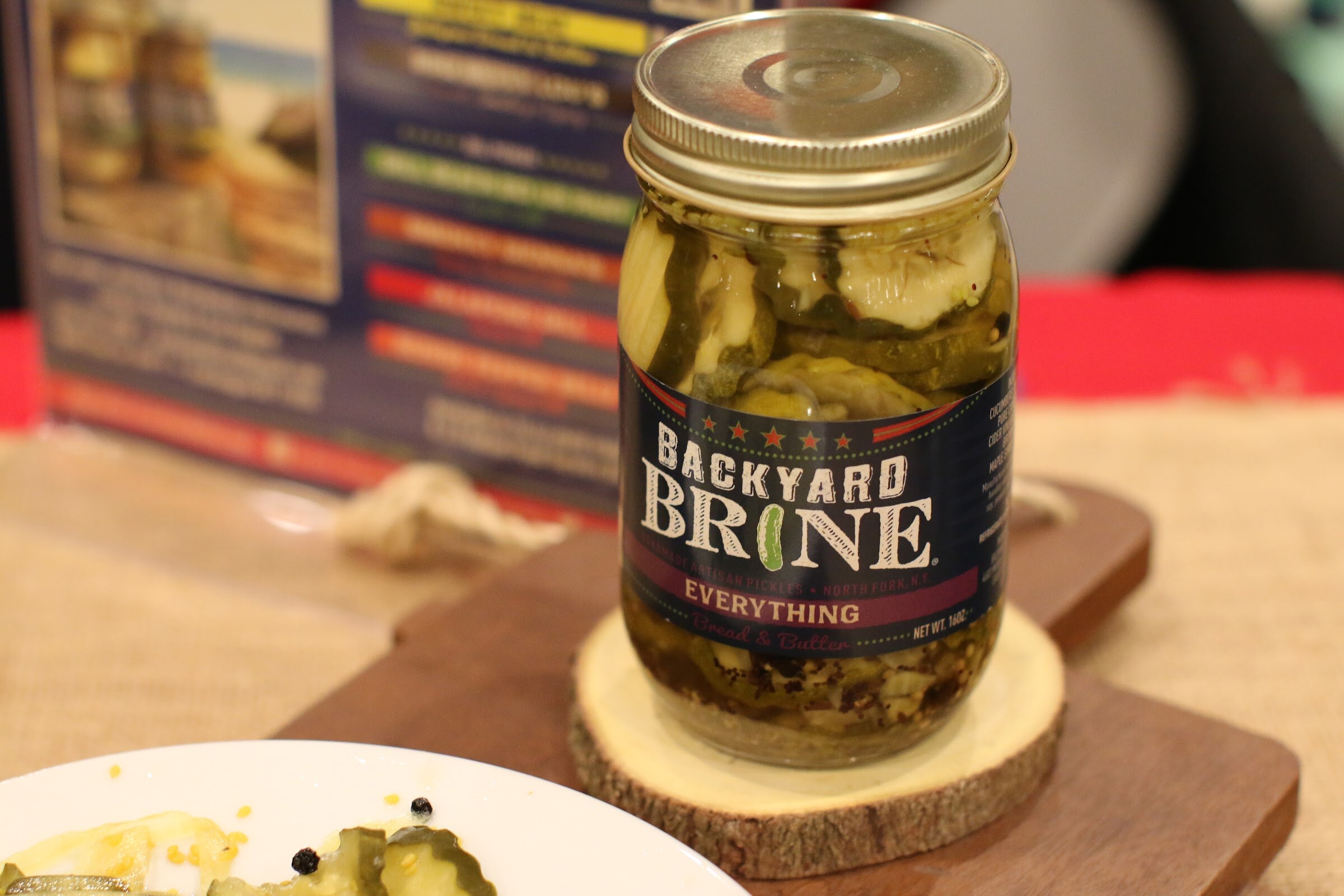 Single jar of Backyard Brine pickles on a round wood slice and cutting board, with a plate of pickle slices at the lower left.