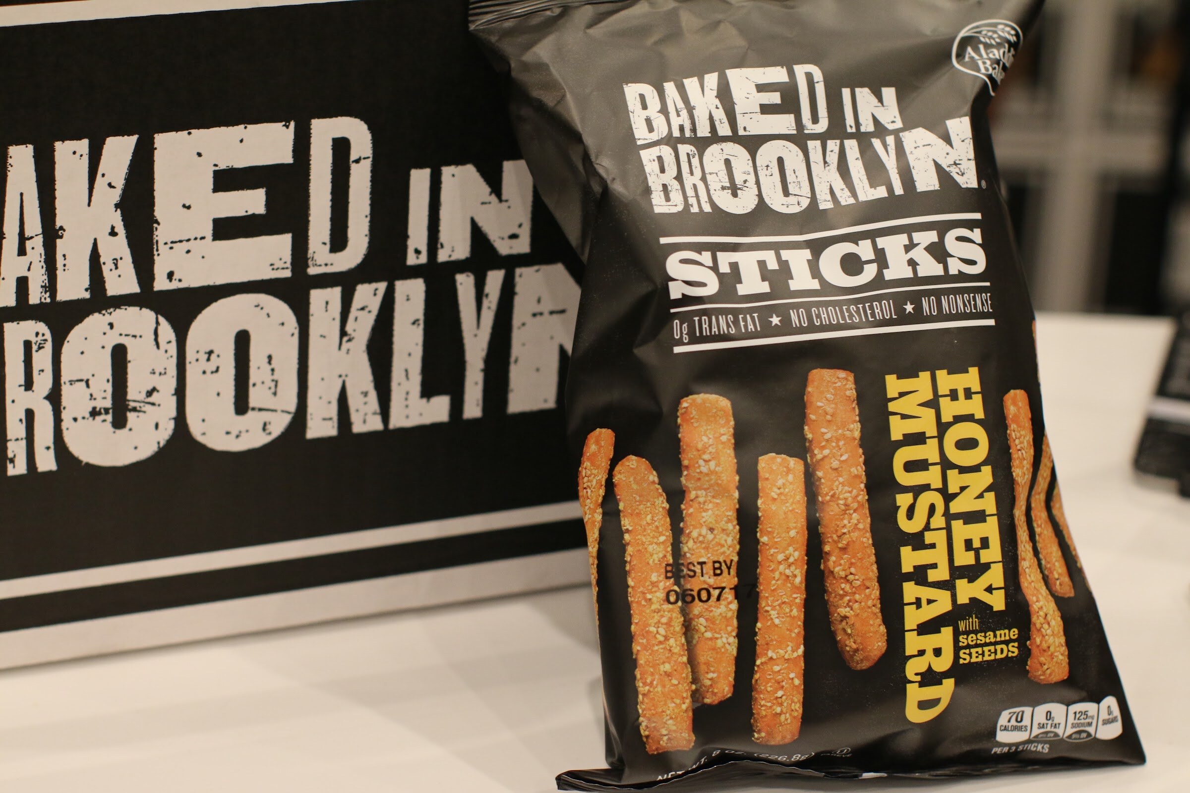 Black bag of Baked in Brooklyn sticks, honey mustard, in front of a large black sign with the brand name.