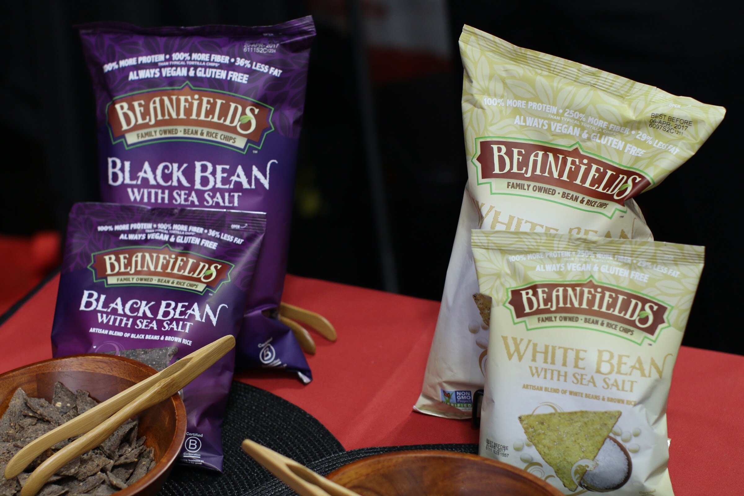 Purple and cream Beanfields chip bags on a red tablecloth beside wooden bowls with serving tongs.