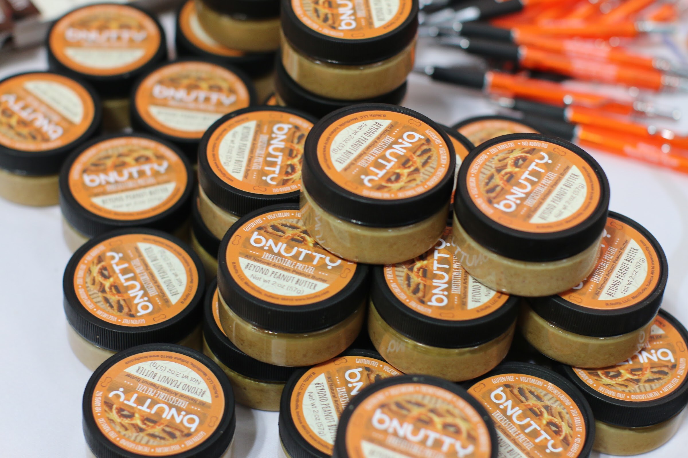 Pile of small bNutty peanut butter jars with orange pretzel-pattern lids on a white table.