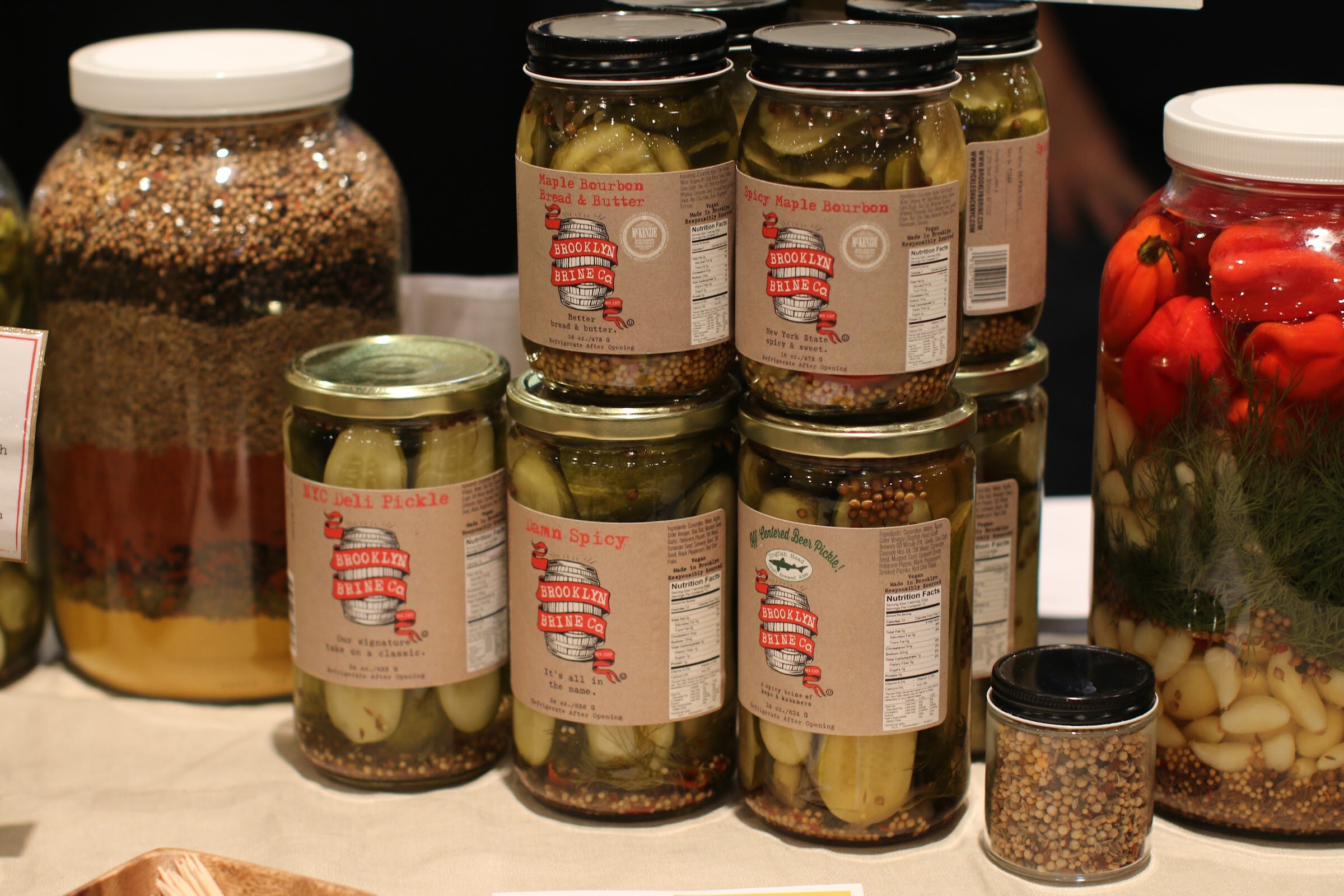 Stacked jars of Brooklyn Brine pickles with kraft labels beside large jars of spices and peppers.